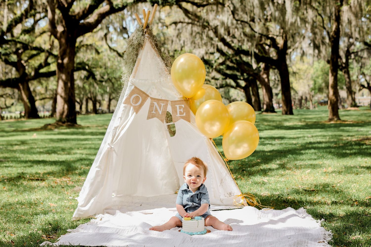 Festive outdoor scene with teepee, yellow balloons and white blanket on grass.