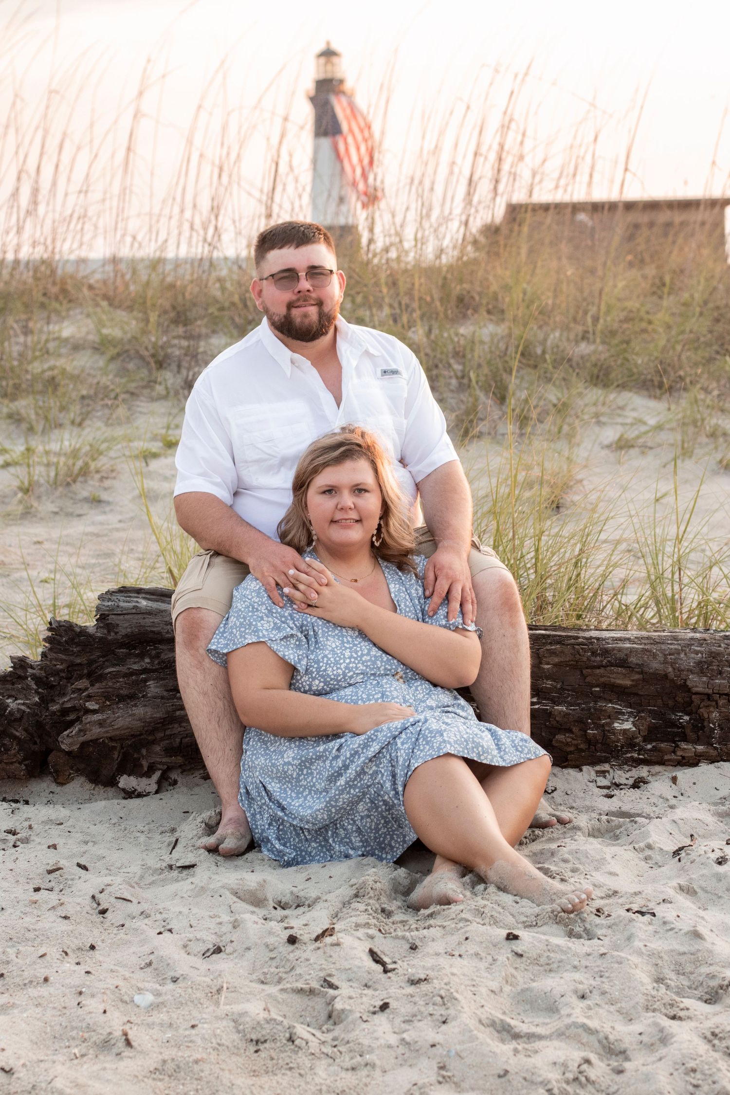 Couple relaxing on sandy beach near driftwood during sunset photoshoot.