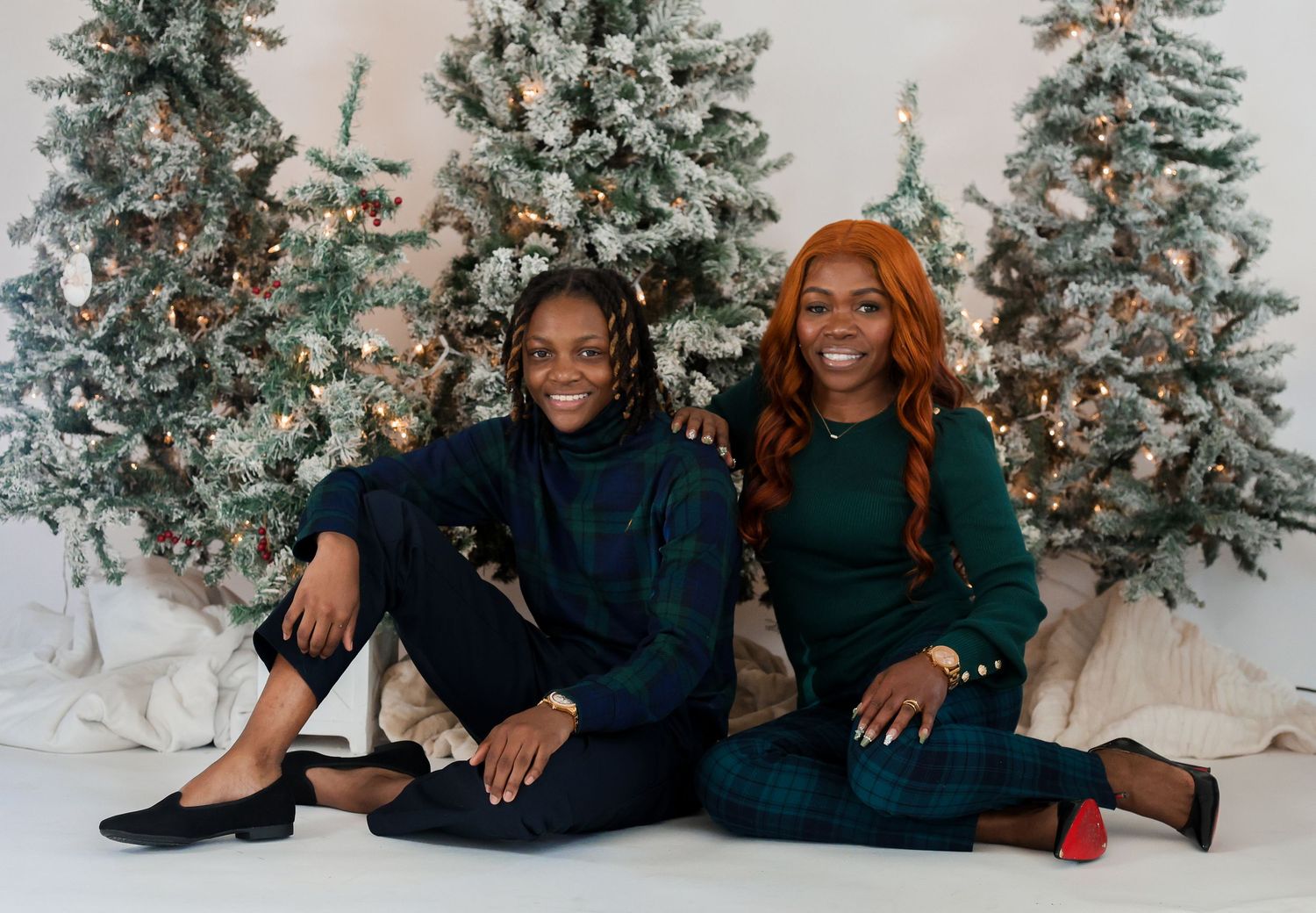 Two people sit together on the floor in front of frosted Christmas trees in a winter-themed photoshoot.