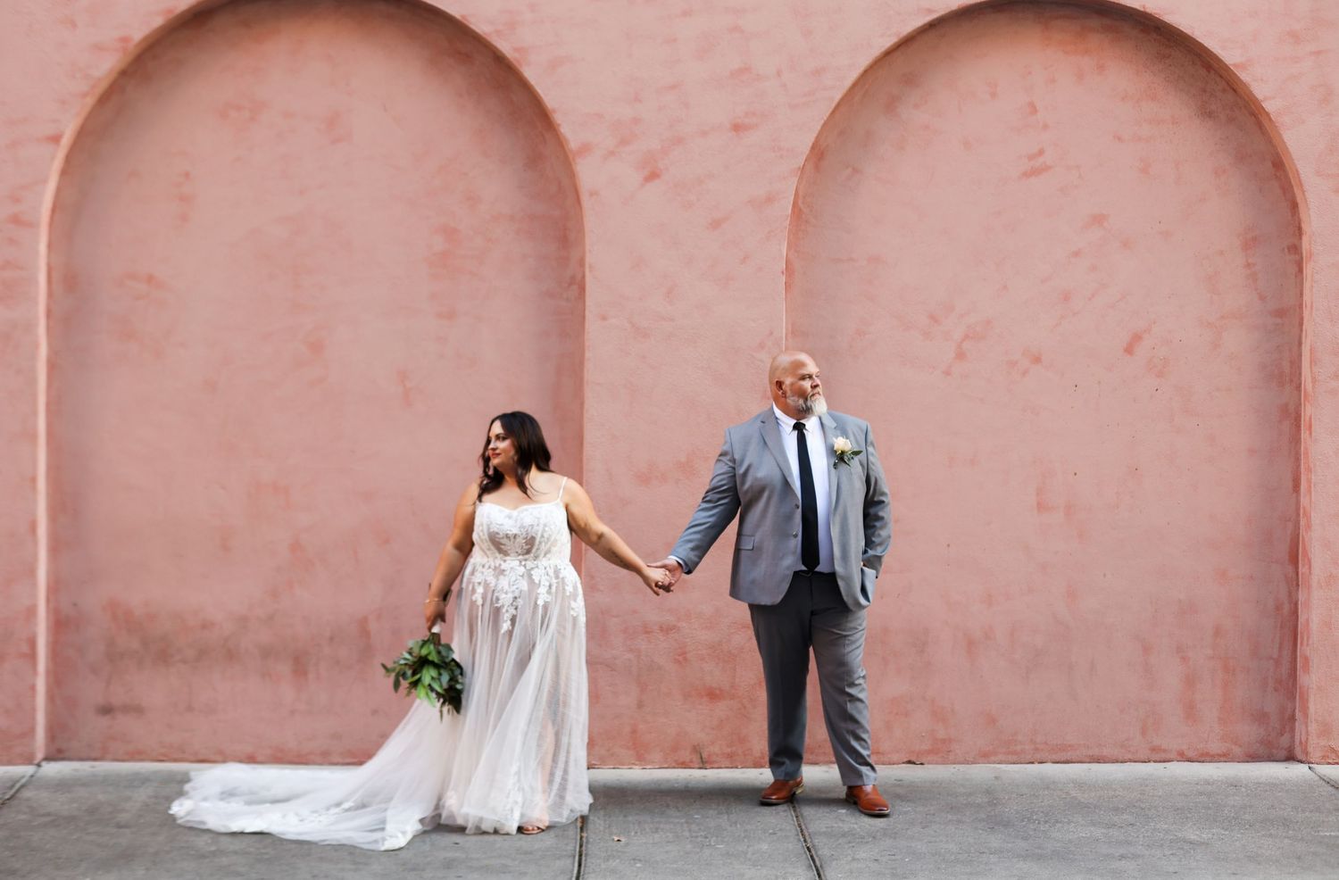 Wedding couple poses against coral colored wall with arched recesses.
