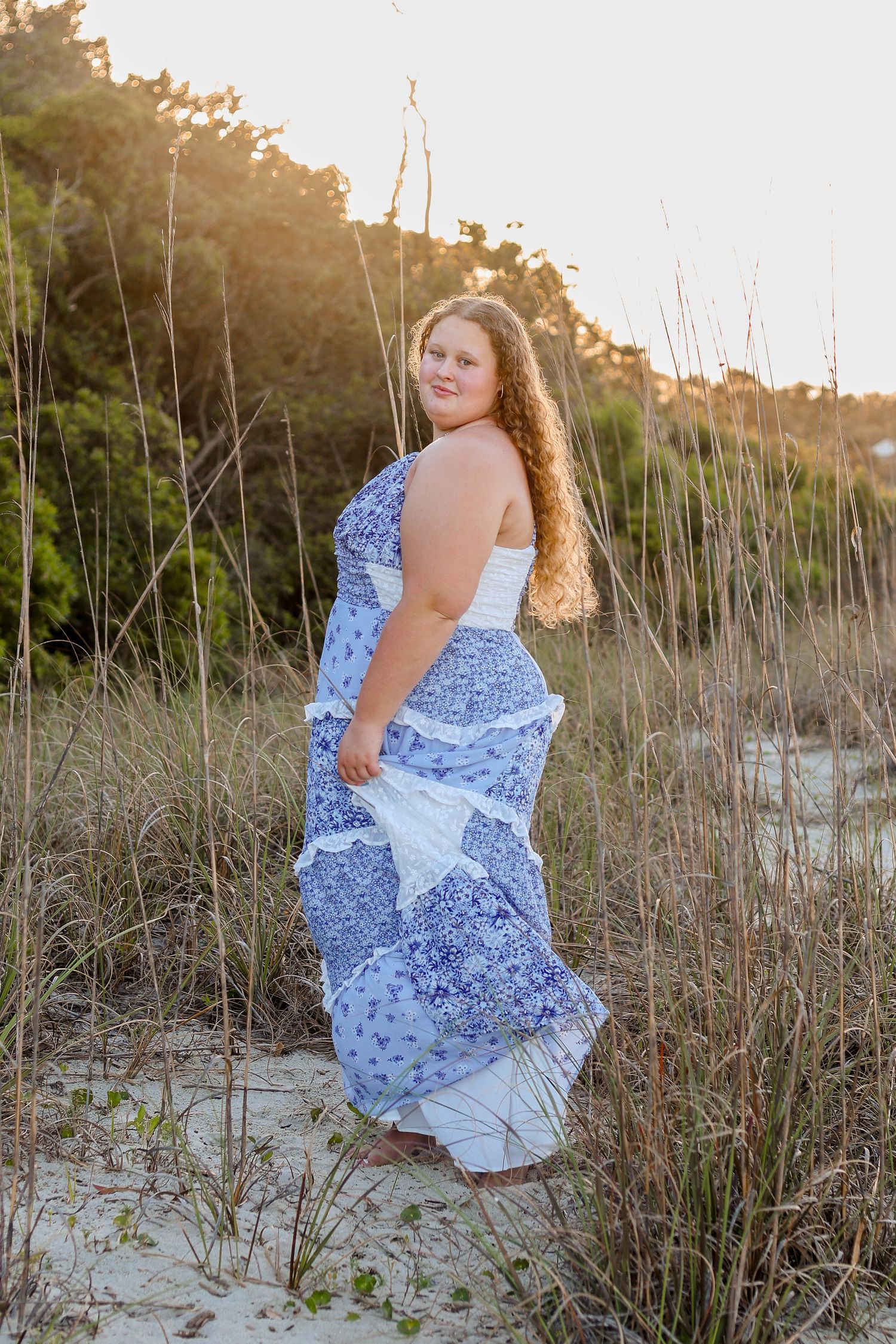 A blue and white floral sundress flows in the coastal breeze at sunset among beach grass and sand dunes.