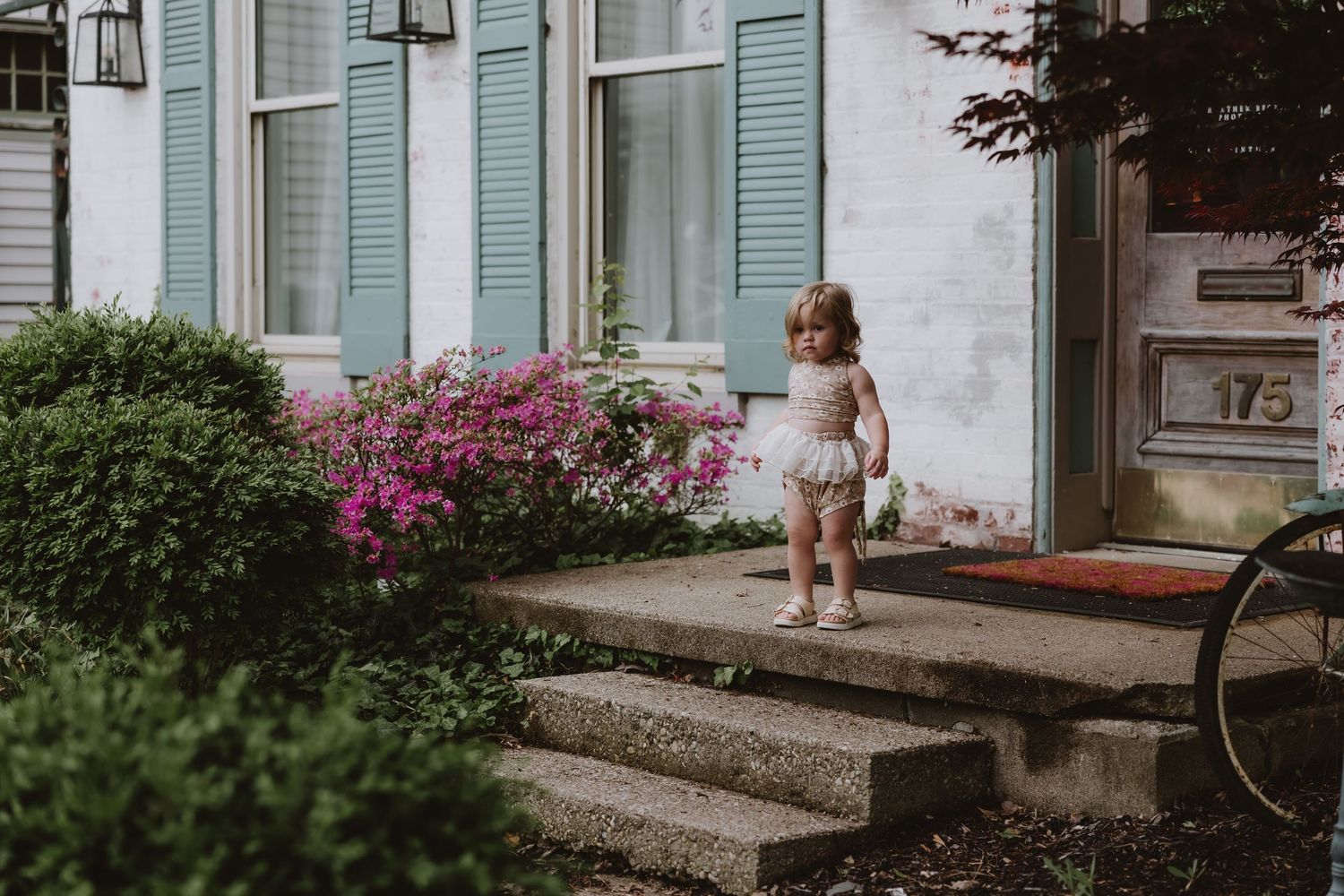 Toddler girl in white dress standing on stone steps outside house number 175 with blue shutters and pink flowers.