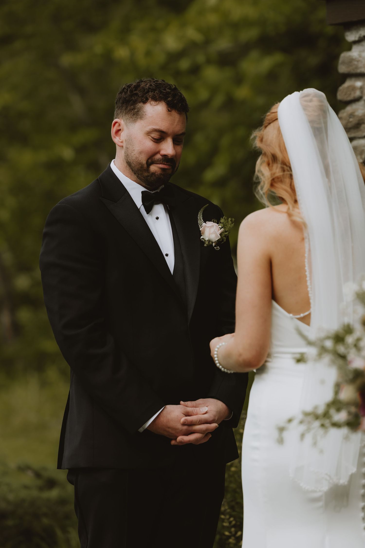 Groom in black tuxedo smiles tenderly at bride in white wedding dress and veil during outdoor ceremony.