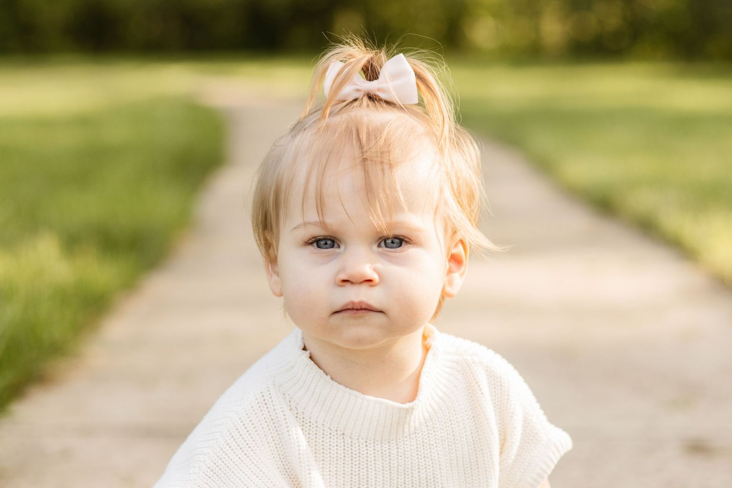 Small child in white sweater stands on pathway with bow in hair.