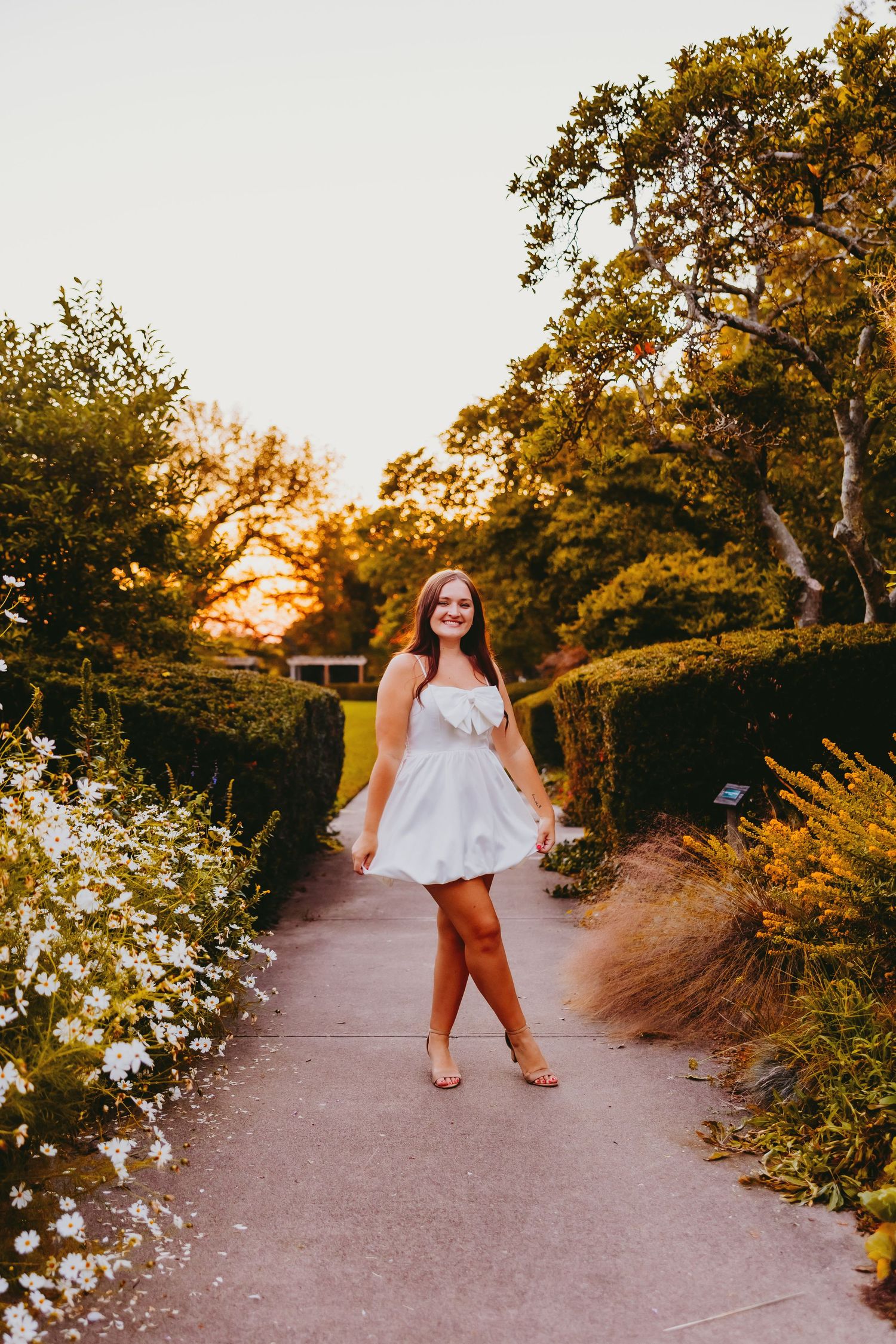 Scenic garden pathway lined with white flowers and trees glowing in golden sunset light.