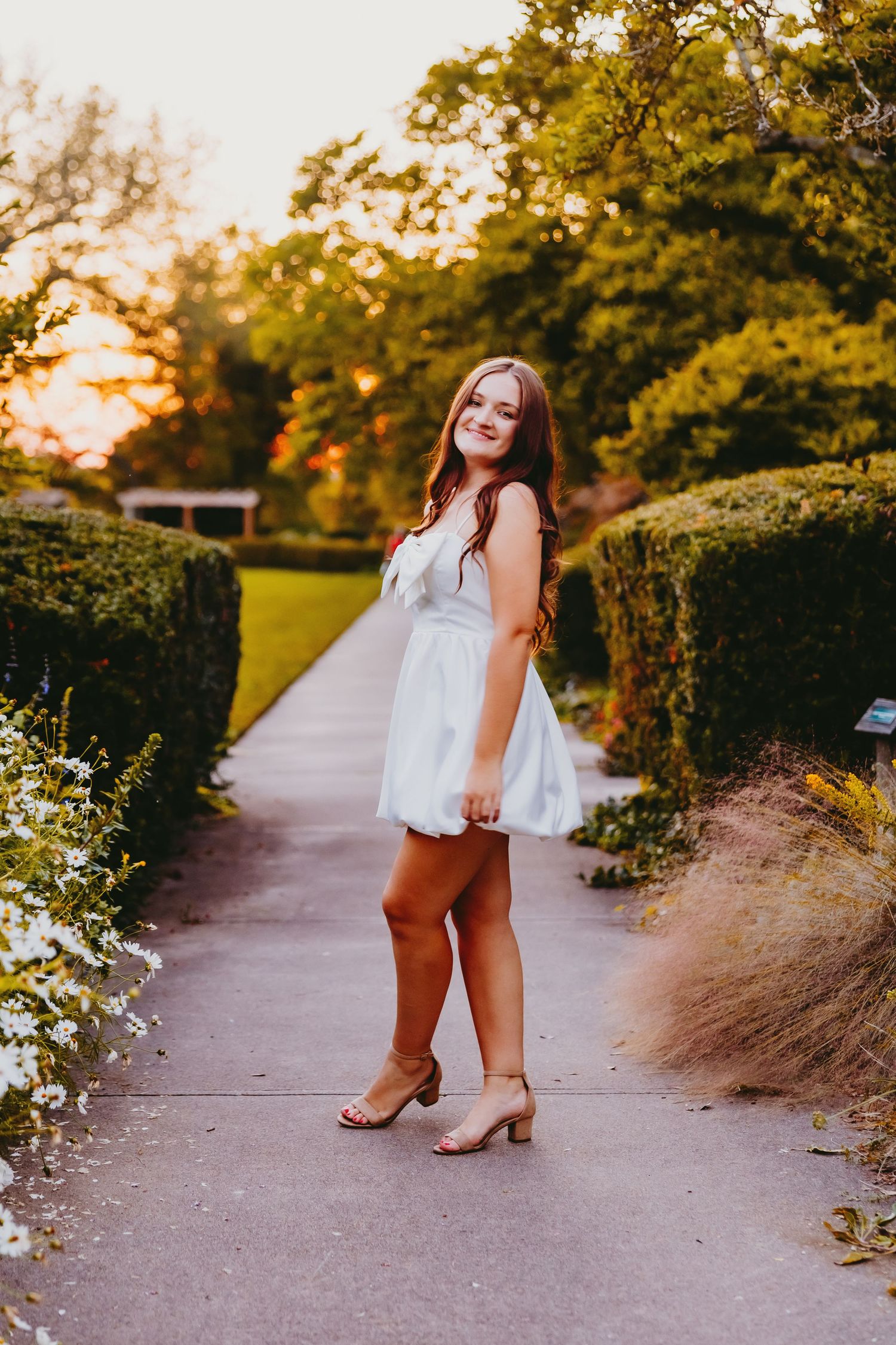 A person in a white top and brown pants poses in a garden path during golden hour sunset lighting.