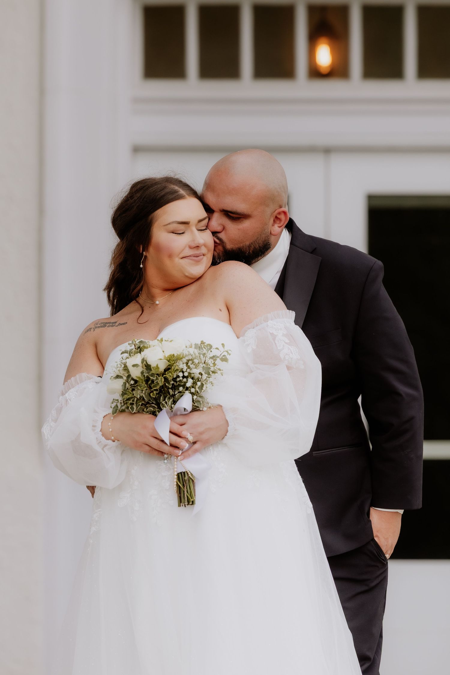 Groom in black tuxedo kissing bride in white off-shoulder gown holding white floral bouquet outdoors.