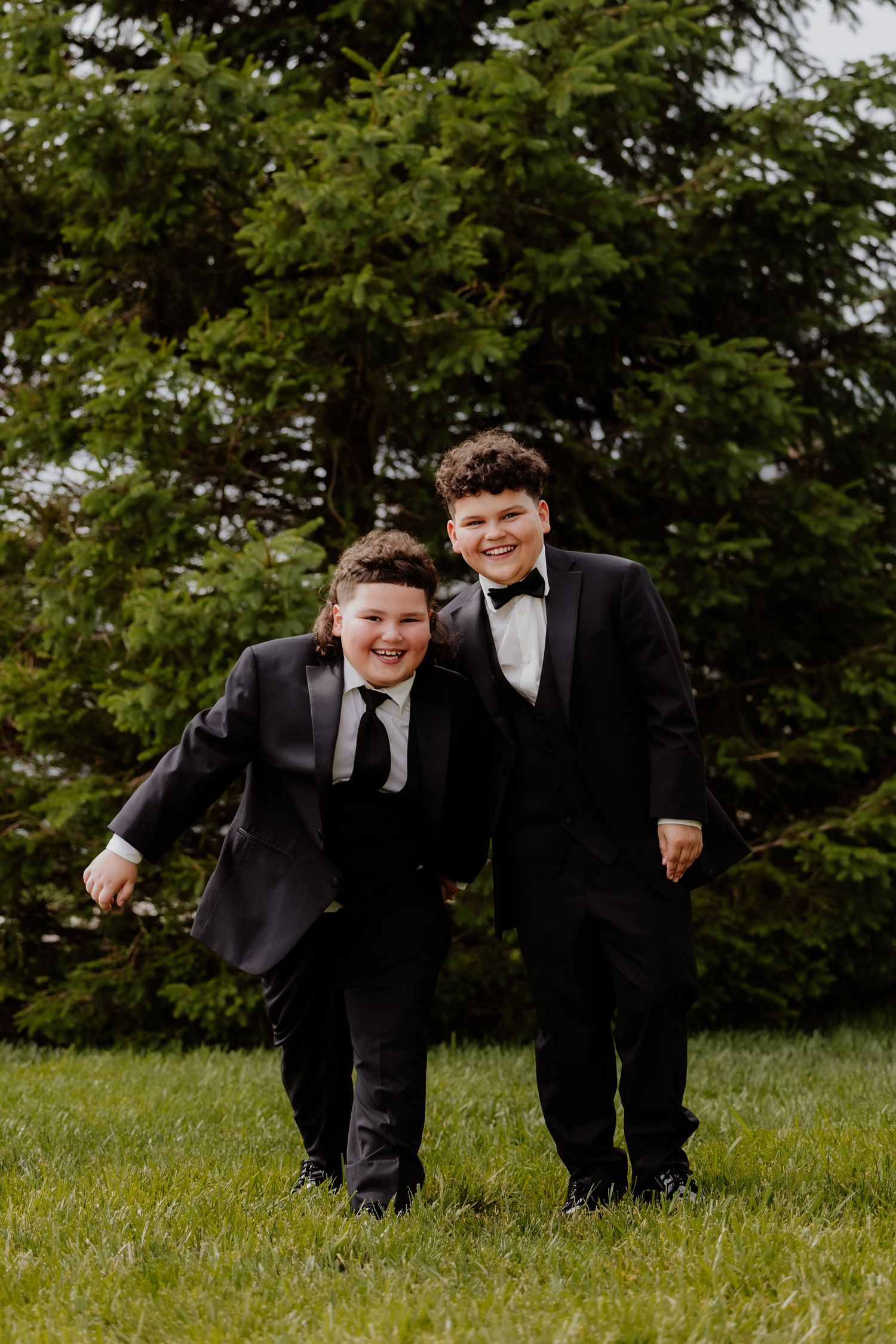 Two smiling boys in black tuxedos stand together on green grass with evergreen trees behind them.