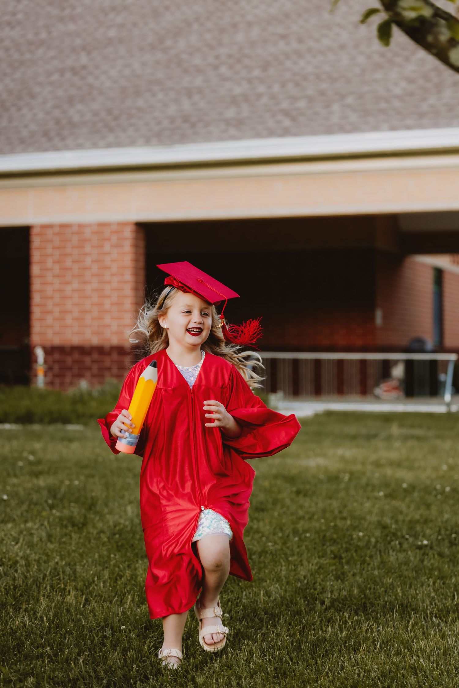 A joyful young girl in a red graduation cap and gown runs across green grass holding a diploma.