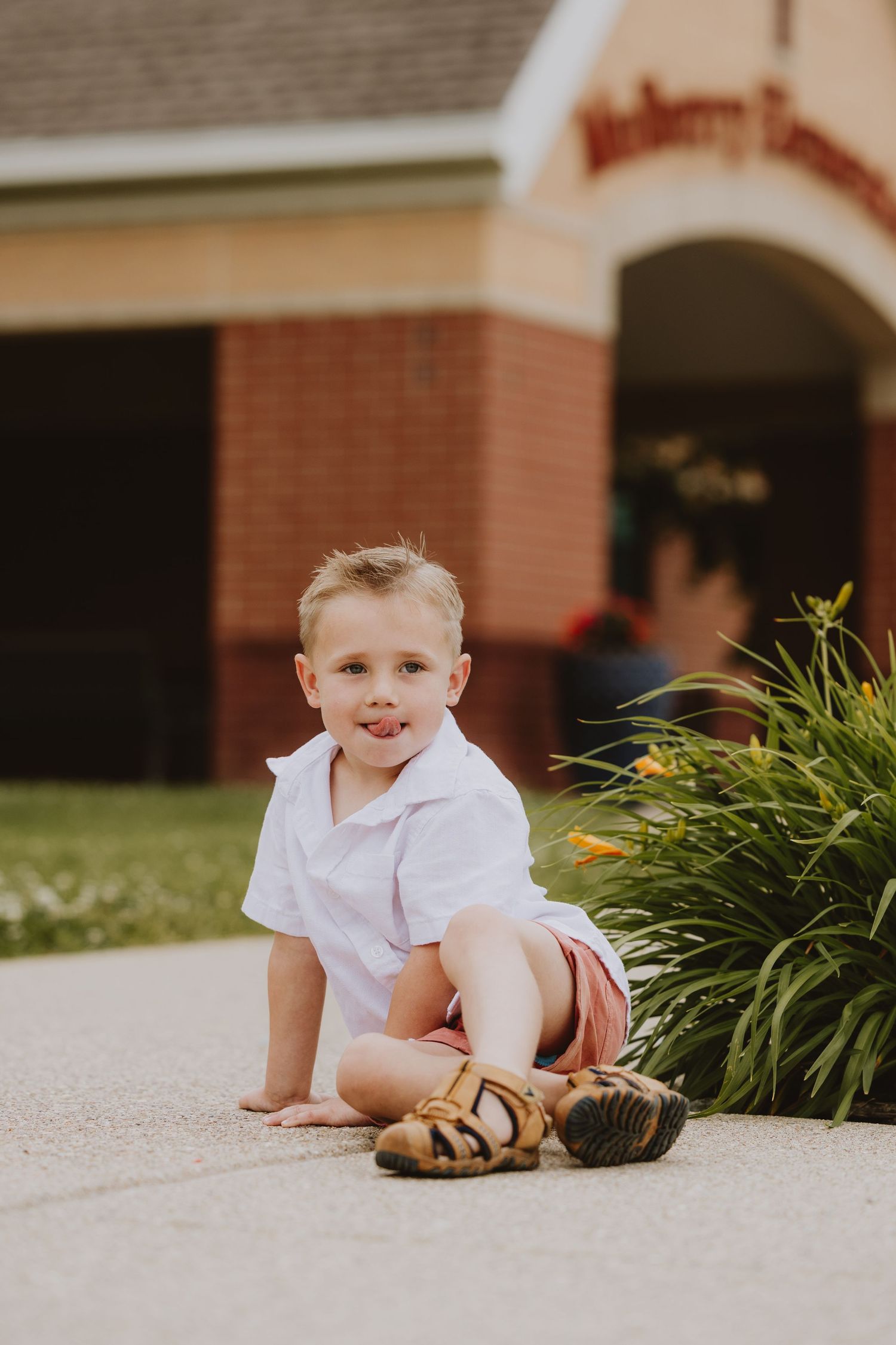 Young blonde boy in white shirt and pink shorts sitting on pavement near flower bed outside brick elementary school.