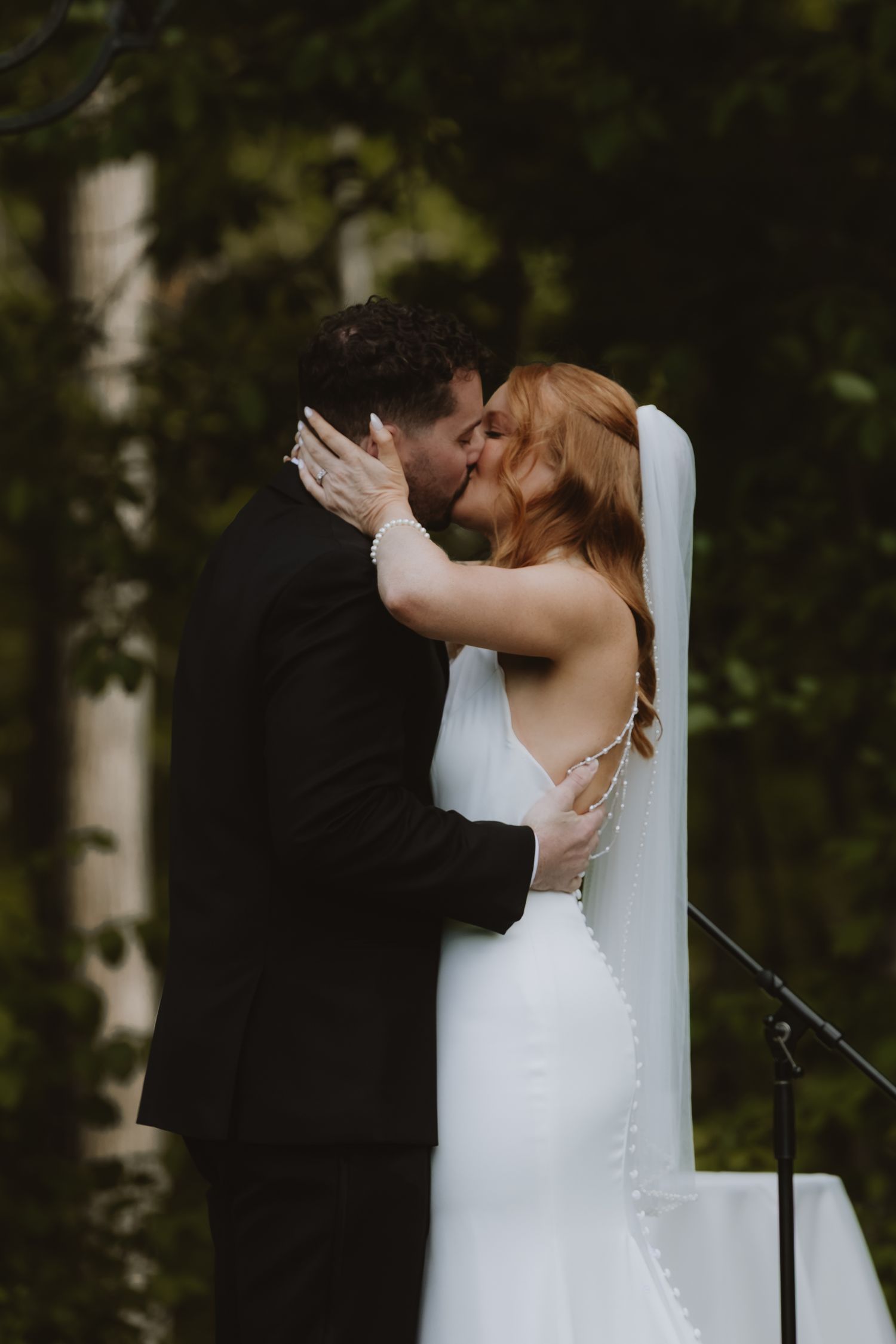 A bride and groom share their first kiss at an outdoor wedding ceremony, surrounded by lush green trees.