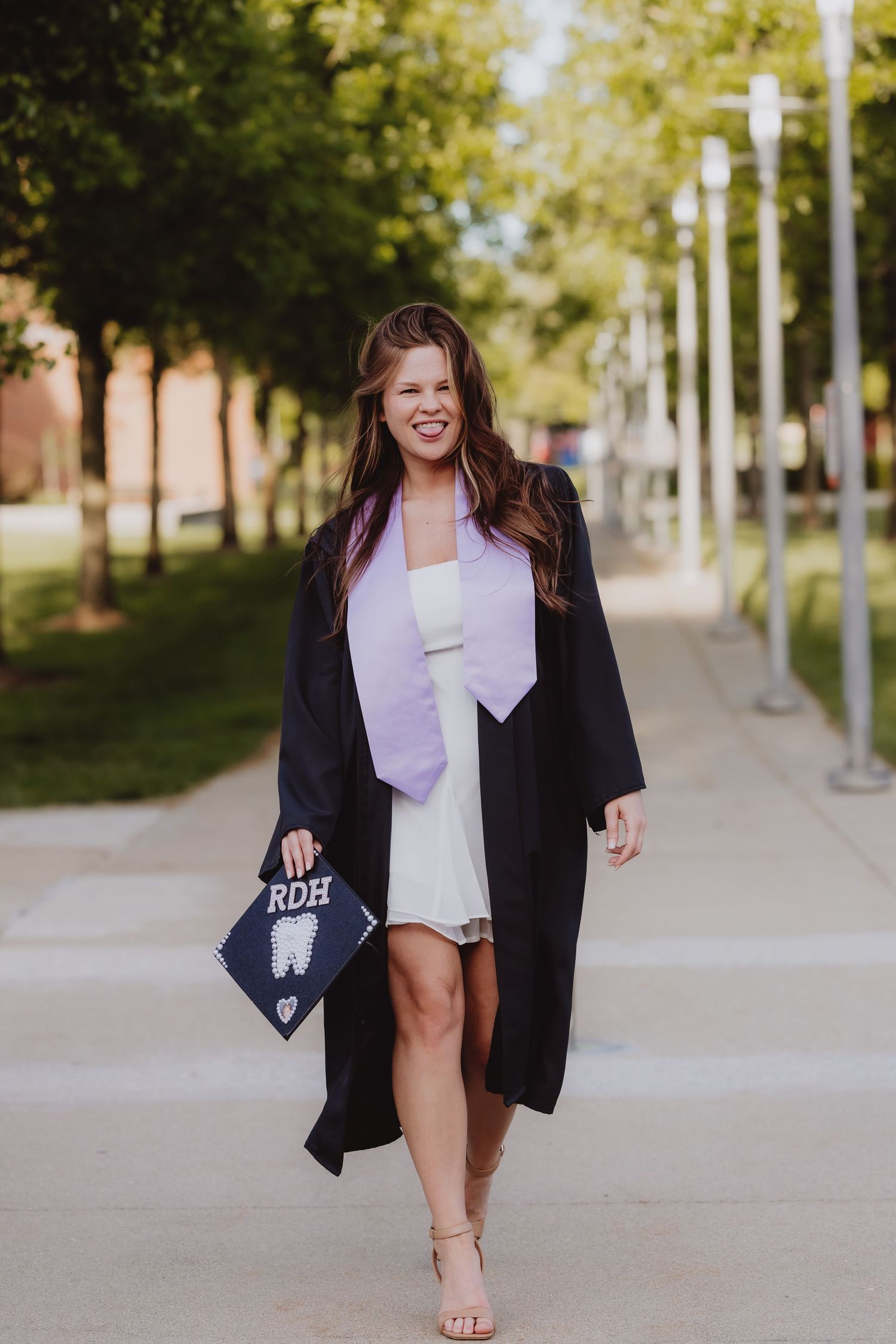 Smiling female graduate in black gown holding RDH cap walks outdoors on campus pathway.