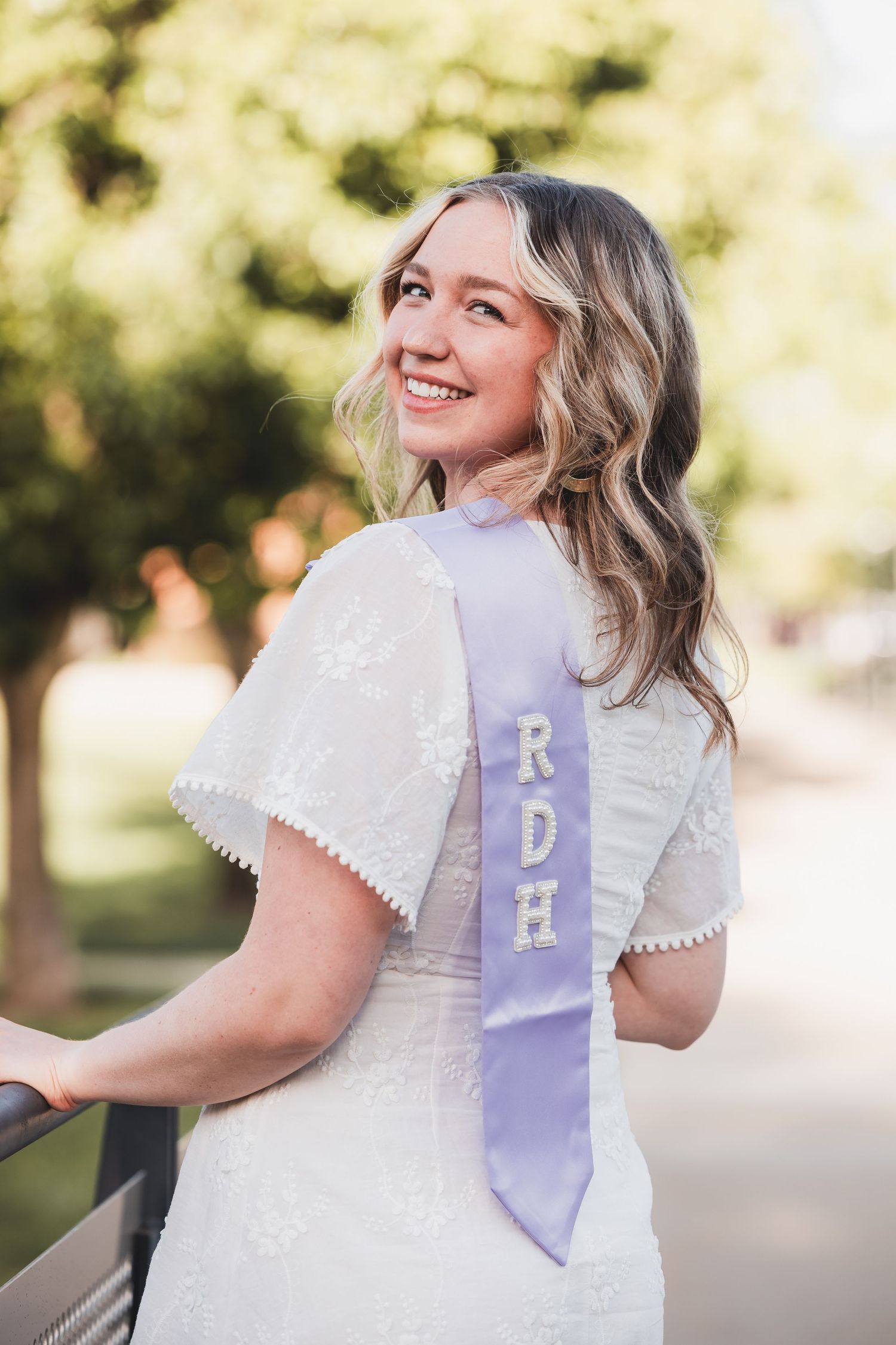 Smiling young woman wearing a white lace dress and lavender RDH graduation sash, posing outdoors.
