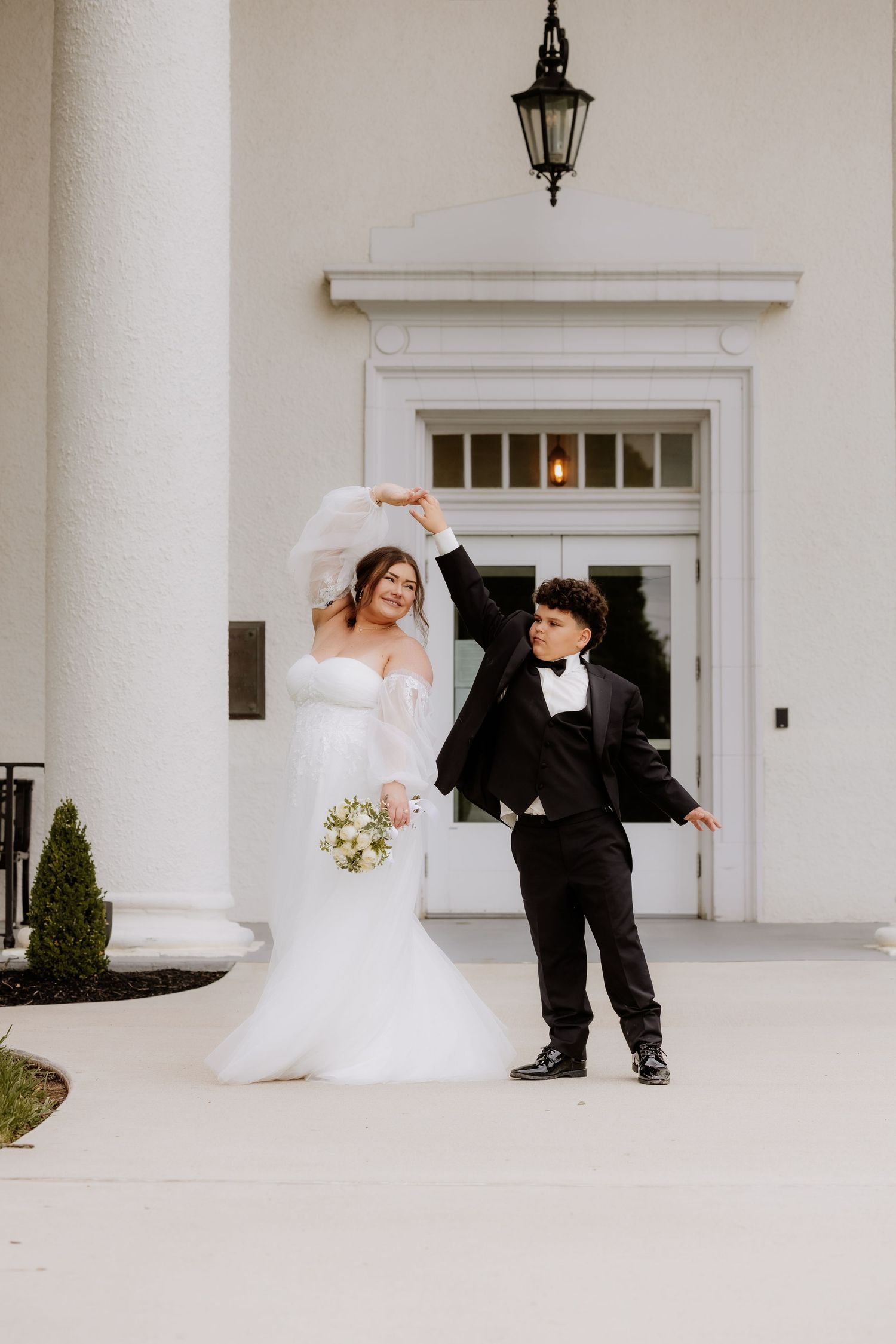Bride in white gown and groom in black tuxedo dancing joyfully outside elegant white venue on their wedding day.