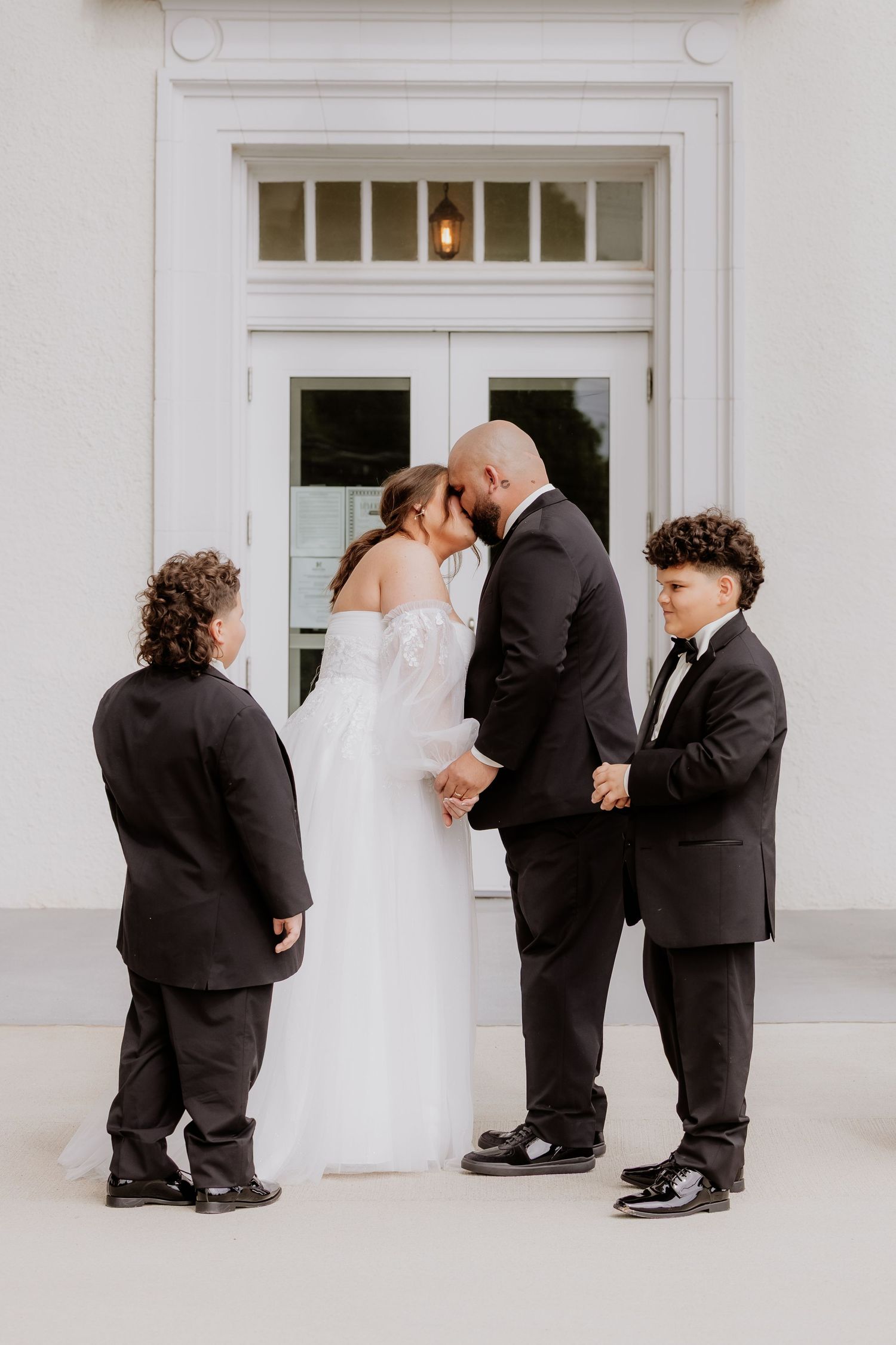 Bride and groom share a kiss flanked by two young boys in tuxedos outside a white building.