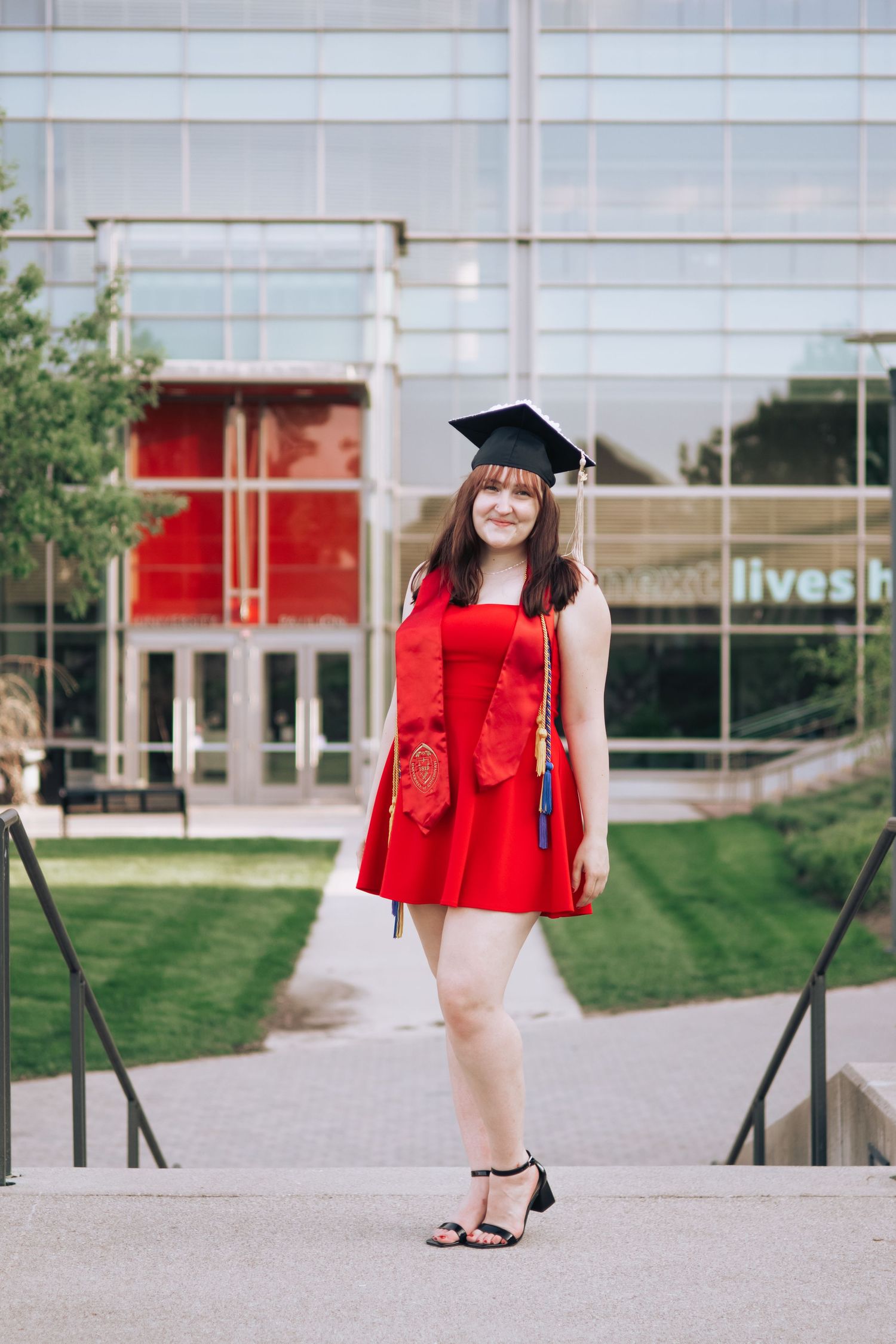A graduate poses on campus steps wearing a red dress and graduation cap.