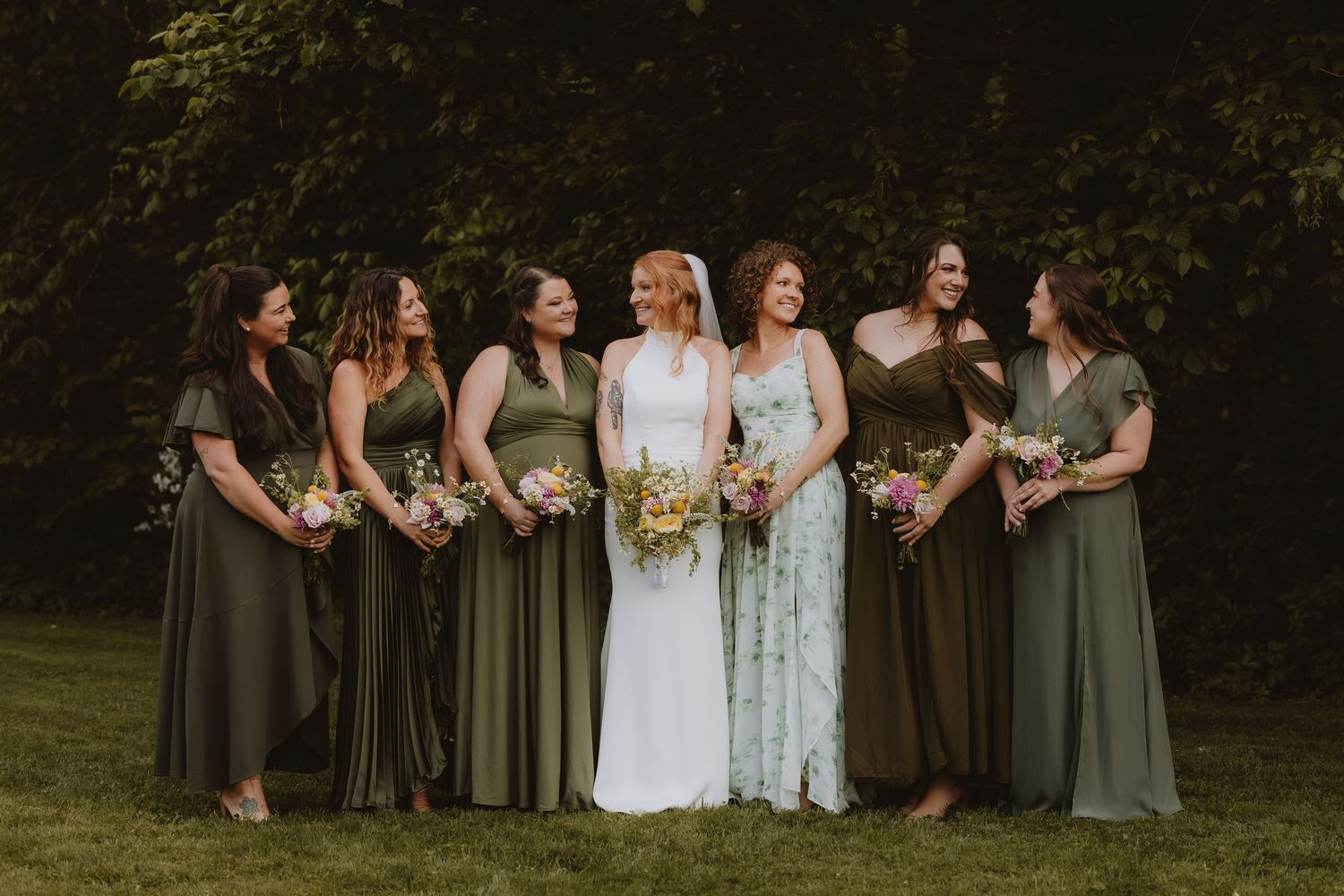 Bride in white floral gown stands with six bridesmaids in mismatched olive green dresses holding bouquets outdoors.