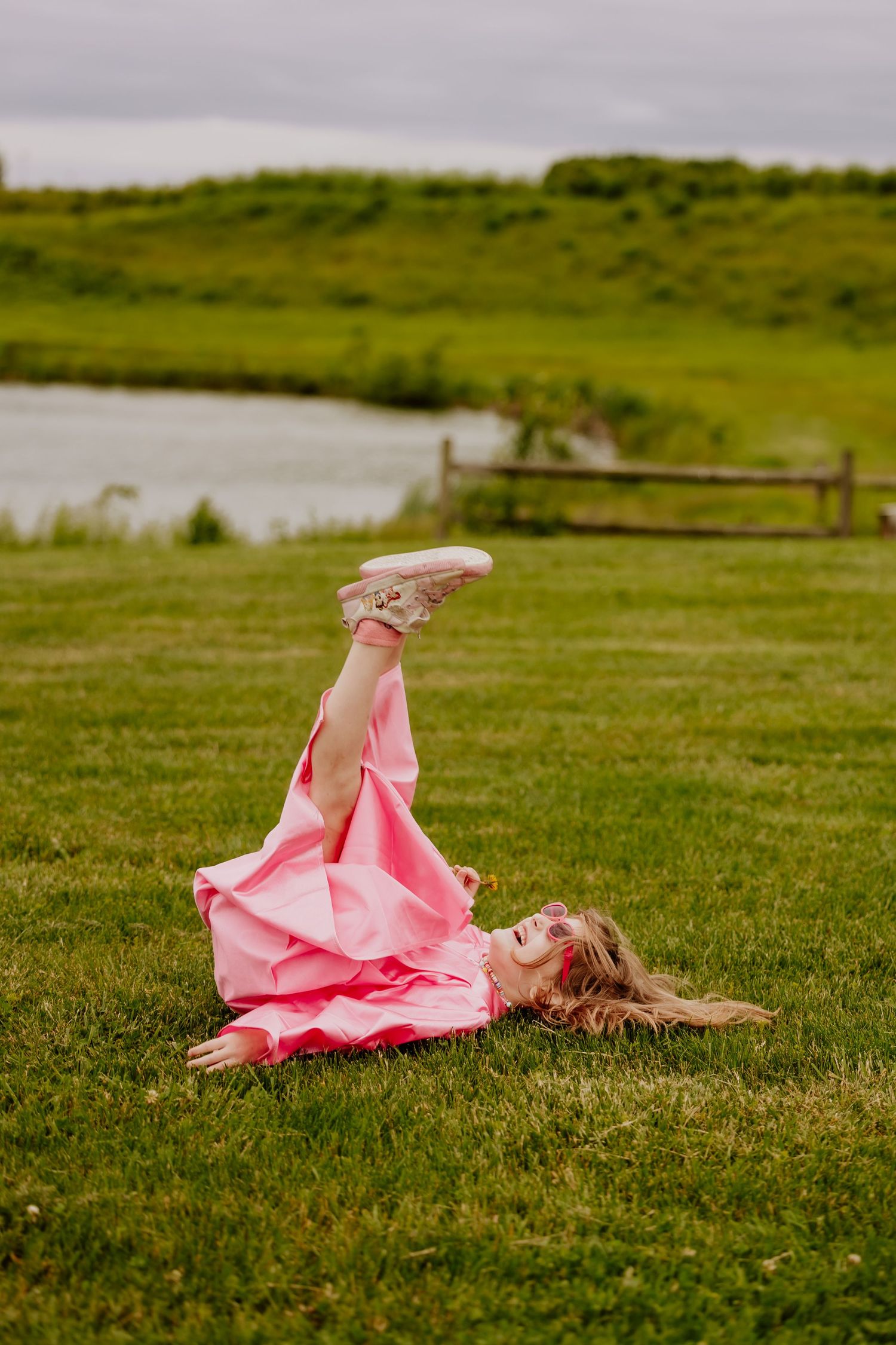 Girl in pink dress lying on green grass with legs up, laughing outdoors near a pond and wooden fence.