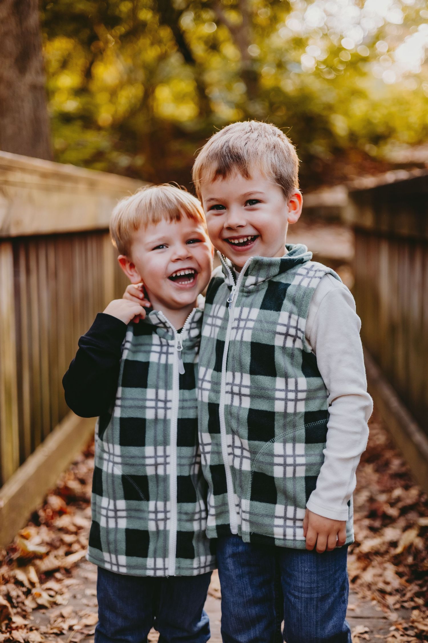 Two young boys in matching plaid vests laugh together on a wooden path surrounded by autumn leaves.