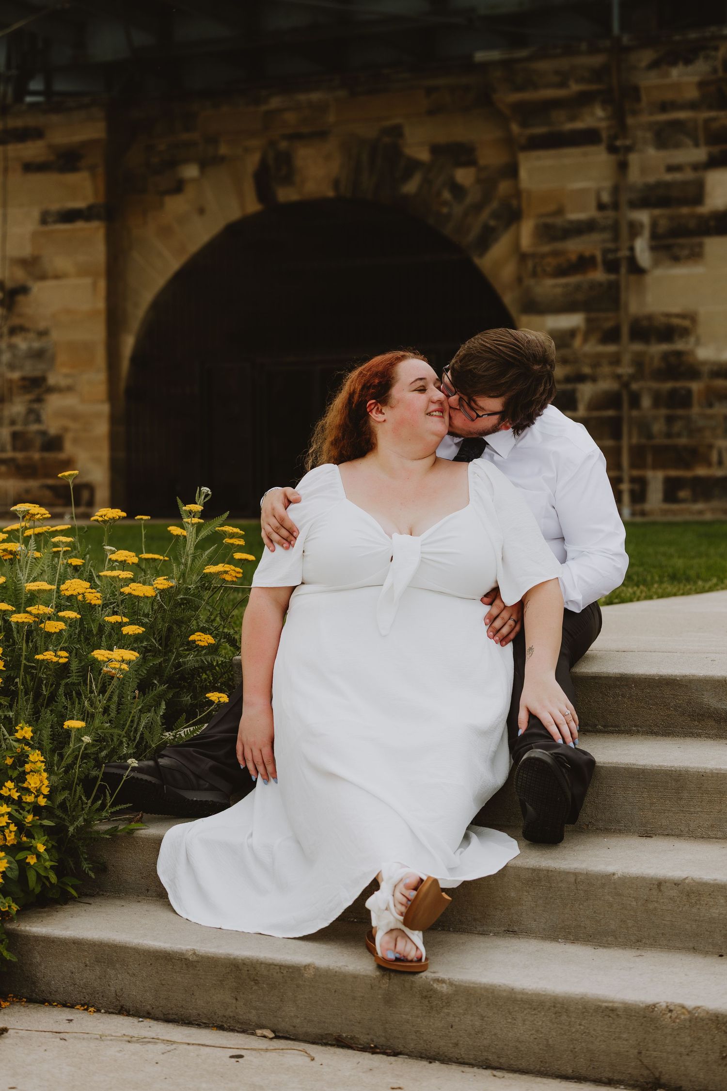 A bride in a white dress sits on stone steps as her groom kisses her cheek beside yellow flowers.