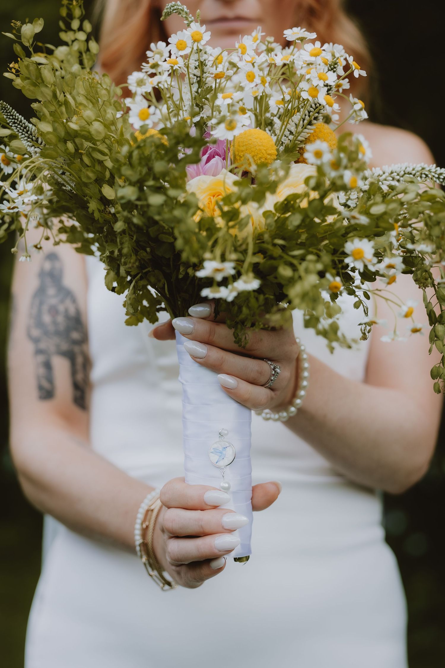 Bride in white dress holding wildflower bouquet with daisies and yellow blooms, wrapped in white ribbon, showing pearl bracelet and tattoo.