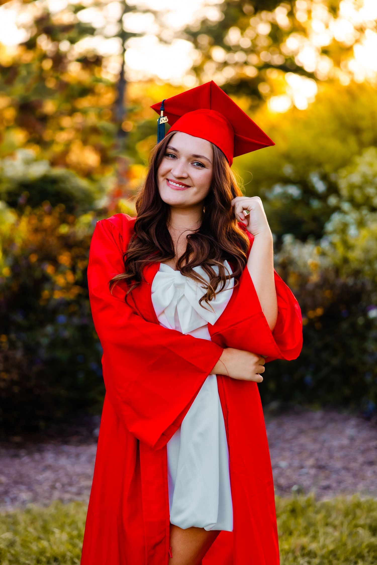 A graduate in bright red cap and gown poses outdoors during golden hour sunset in a white dress.