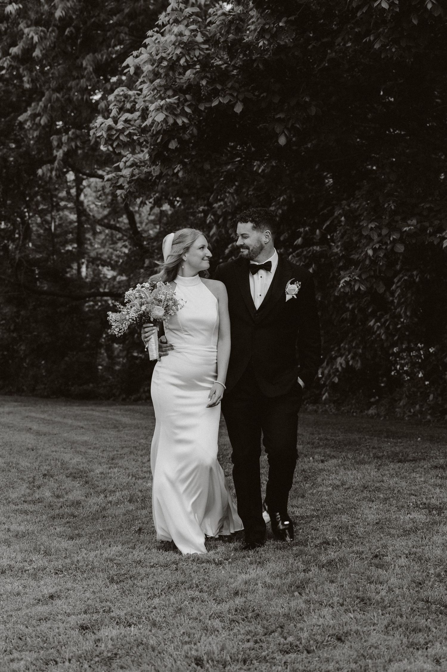 Black and white photo of a bride and groom walking together on a lawn, smiling at each other on their wedding day.