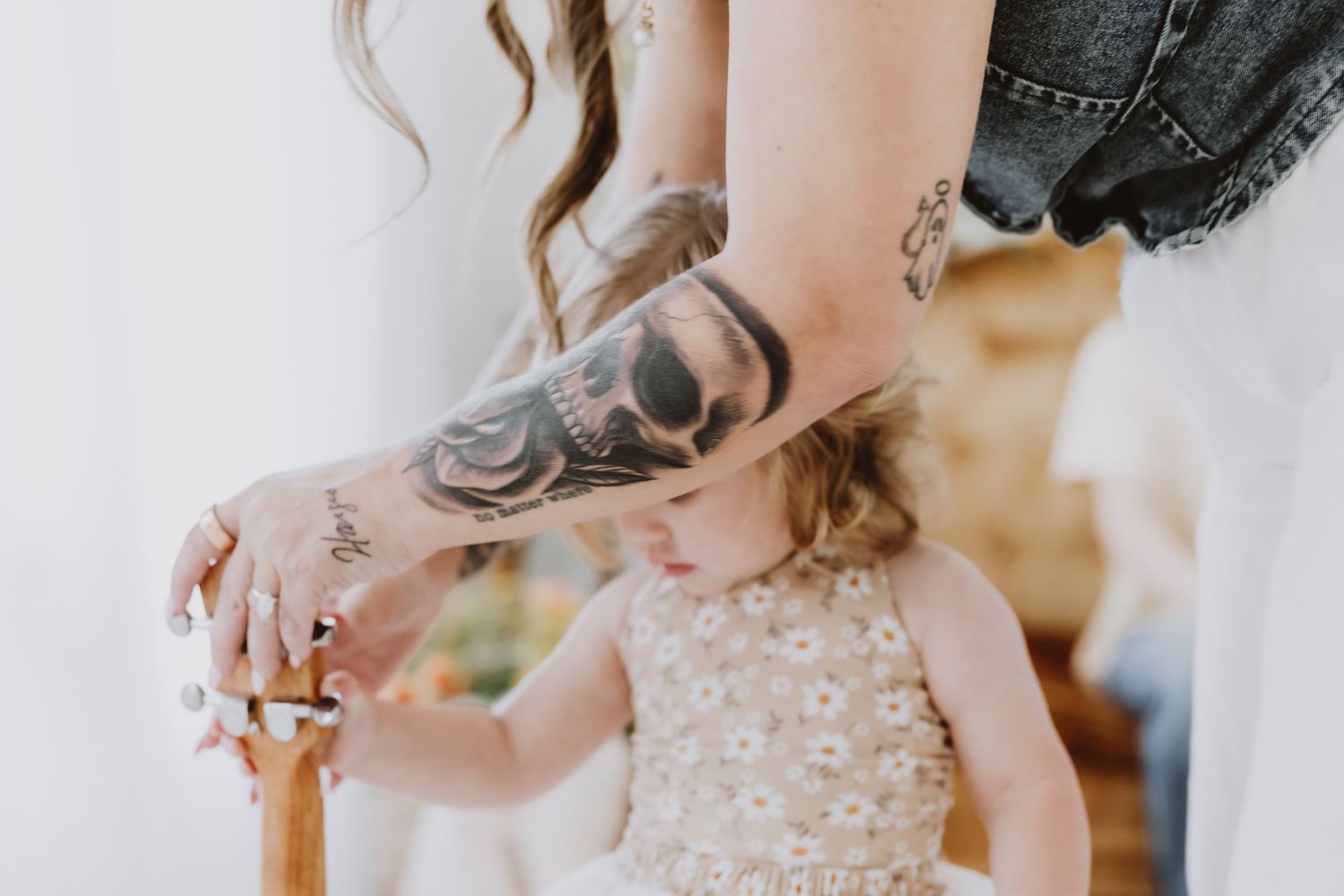 Tattooed arm of a parent resting on a wooden chair beside a young toddler girl in a floral dress.