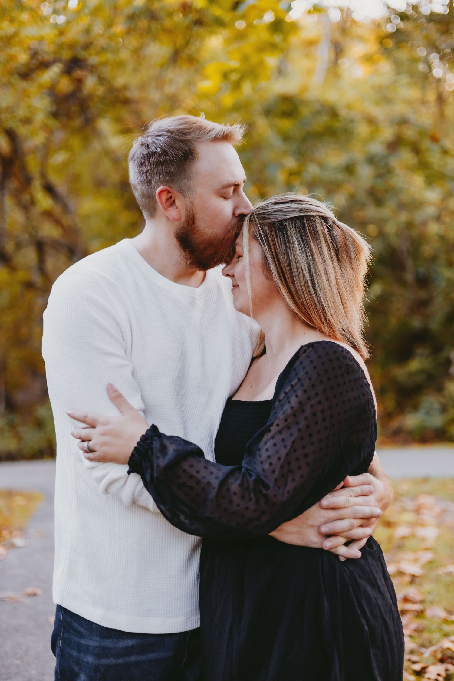 A couple embraces tenderly outdoors in autumn, man kissing woman's forehead, surrounded by golden fall foliage.