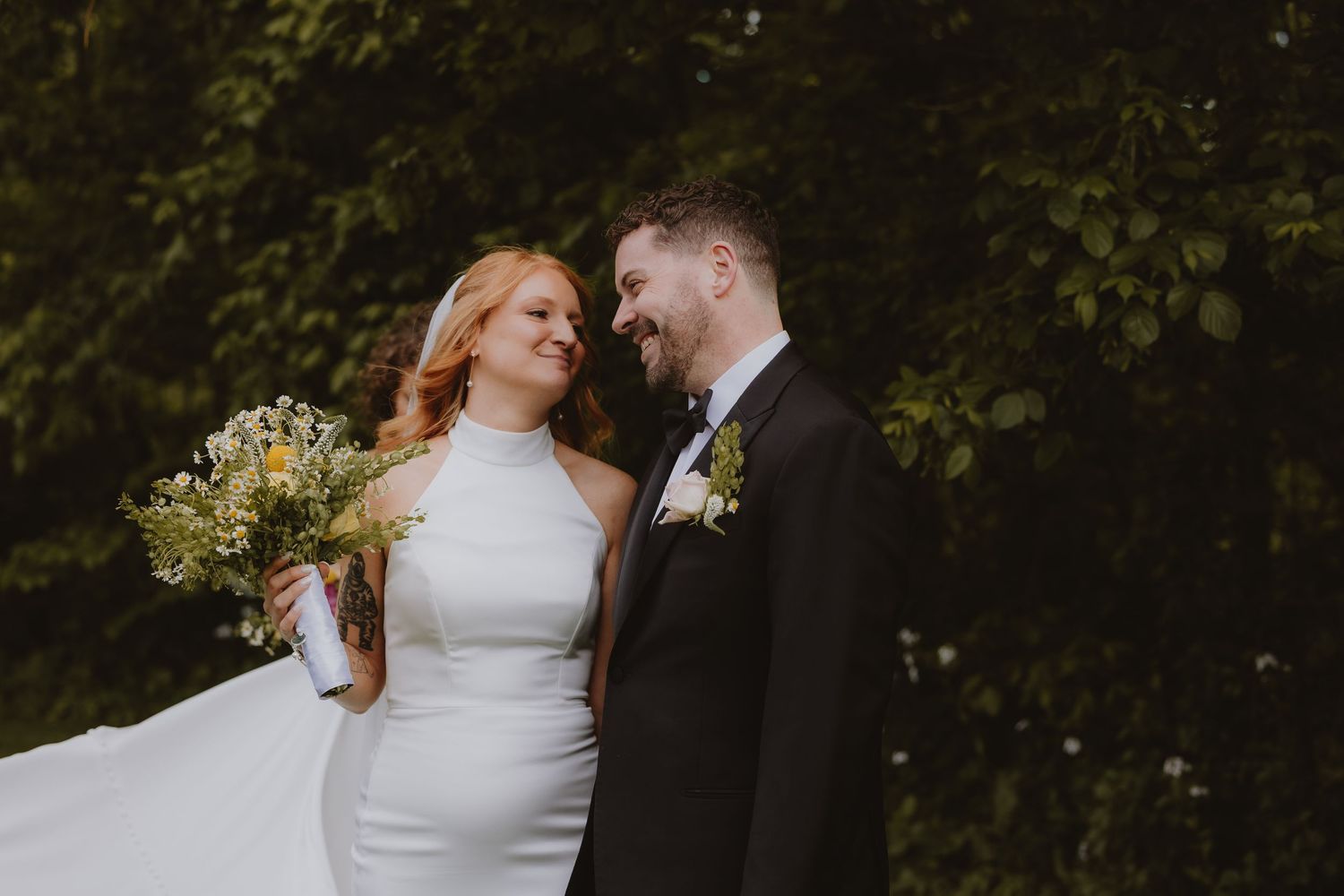Bride and groom share a romantic moment outdoors, she holds a wildflower bouquet in a white halter gown, he wears a black suit.