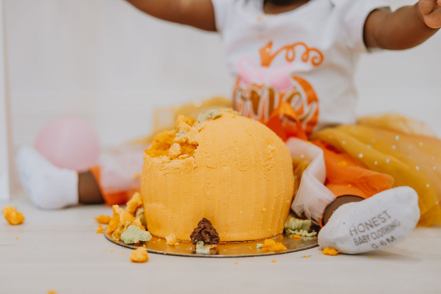 Baby smash cake session with orange frosted cake on silver platter, child in white outfit reaching toward dessert.