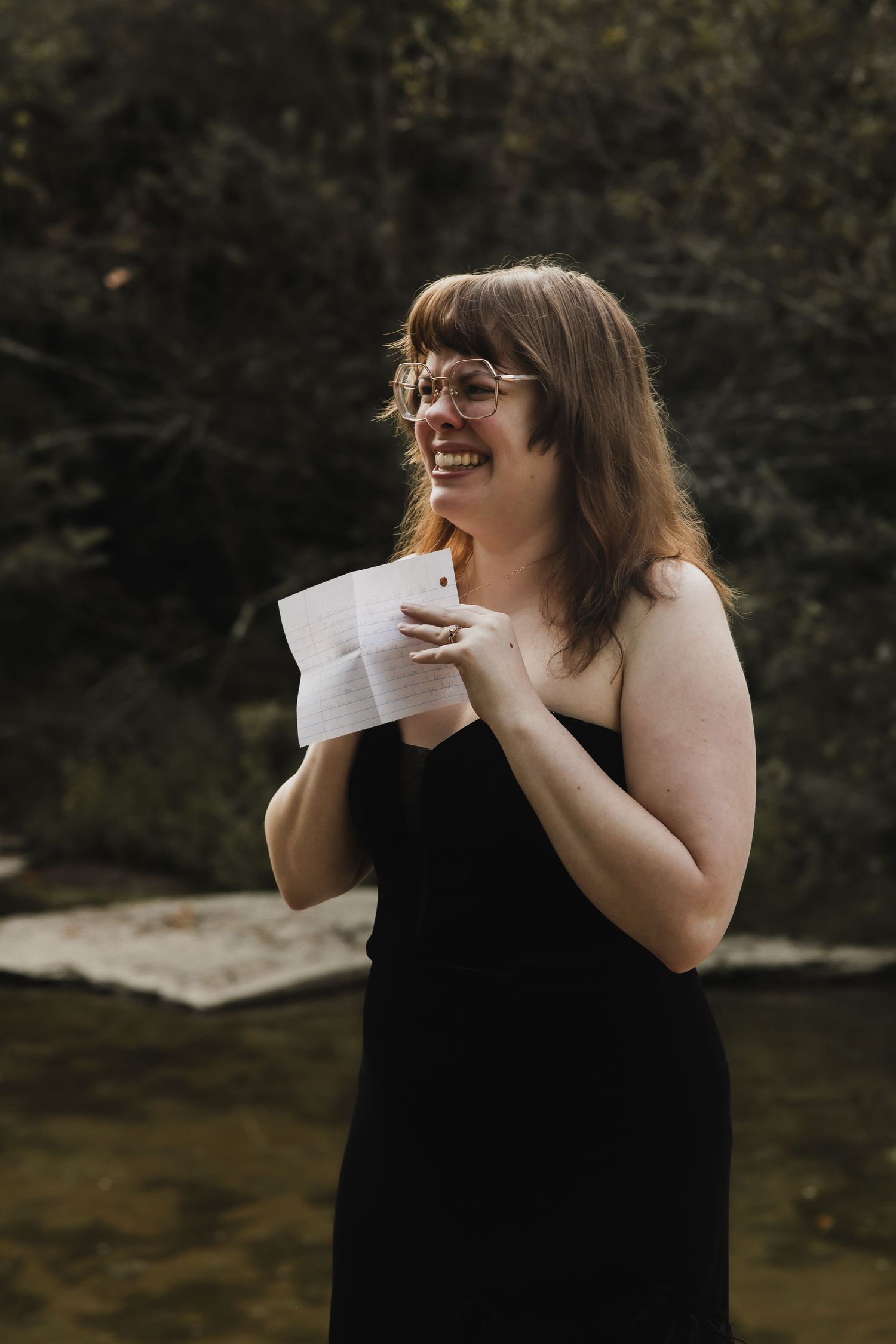 A person in a black tank top laughs joyfully while holding papers outdoors in a natural setting.