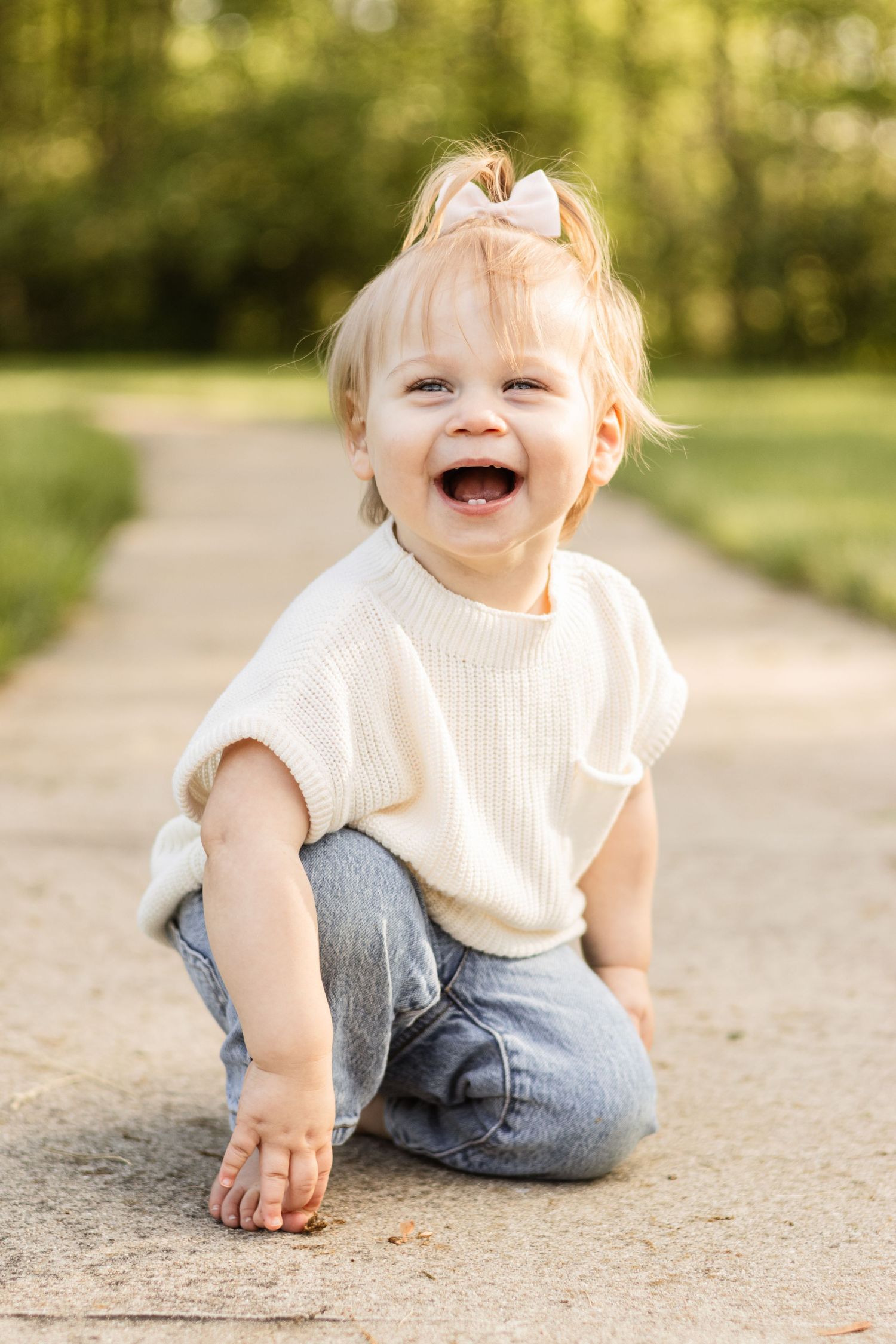 Laughing toddler in white sweater sits on concrete path during outdoor photoshoot.