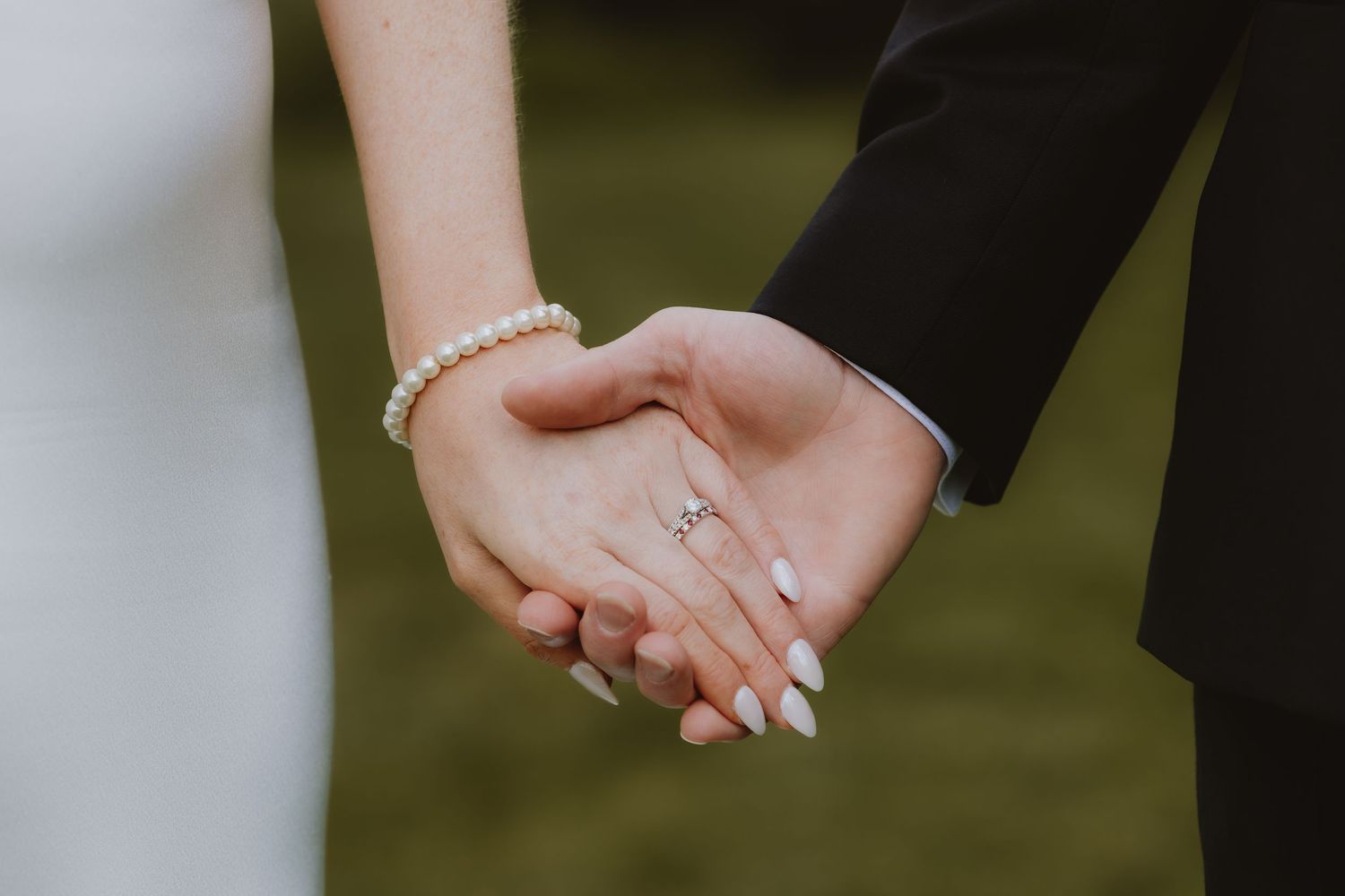 Bride and groom holding hands, showcasing diamond engagement ring, pearl bracelet, and white manicure on wedding day.