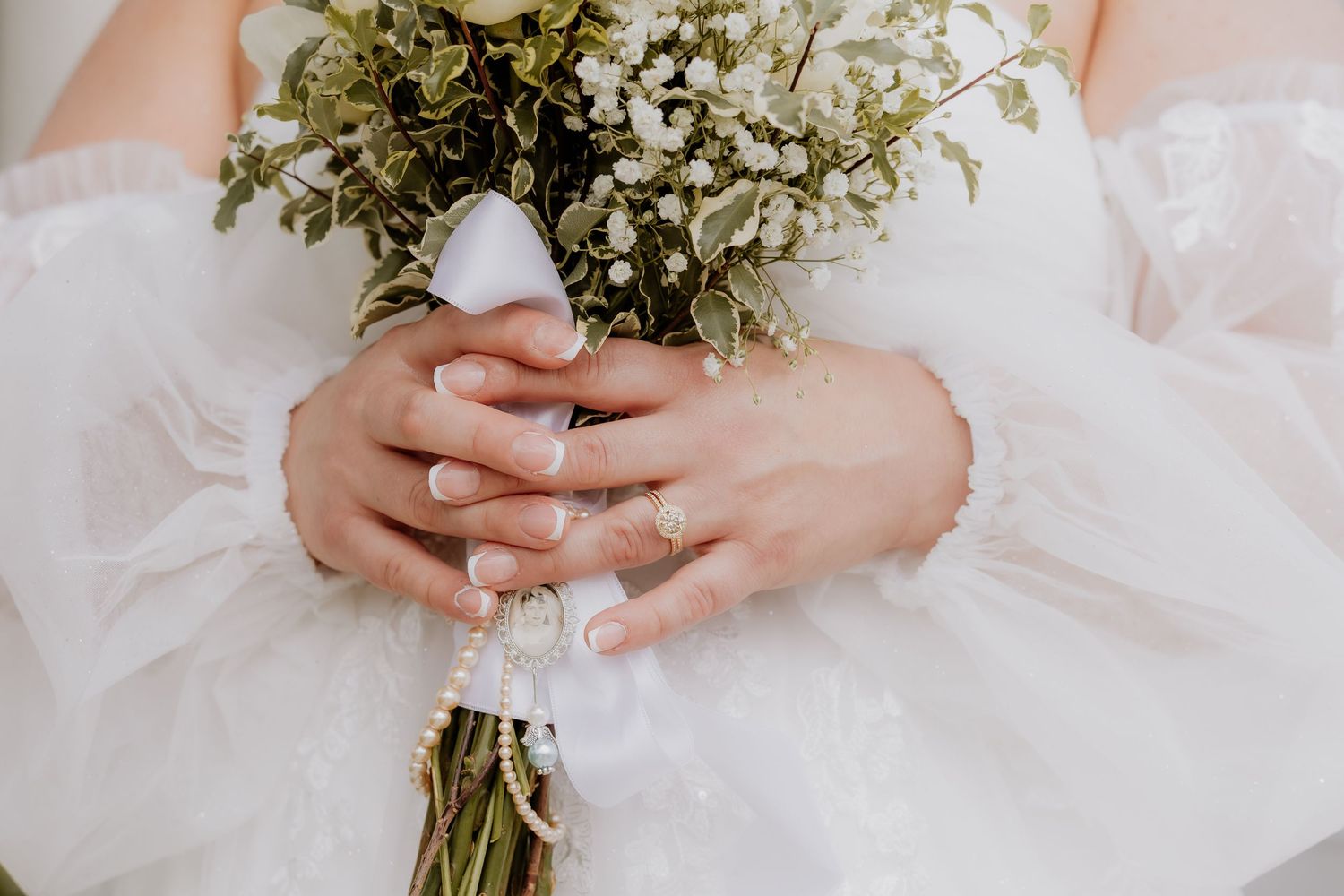 Bride holding a white floral bouquet with greenery, wearing a white dress and delicate rings on her fingers.