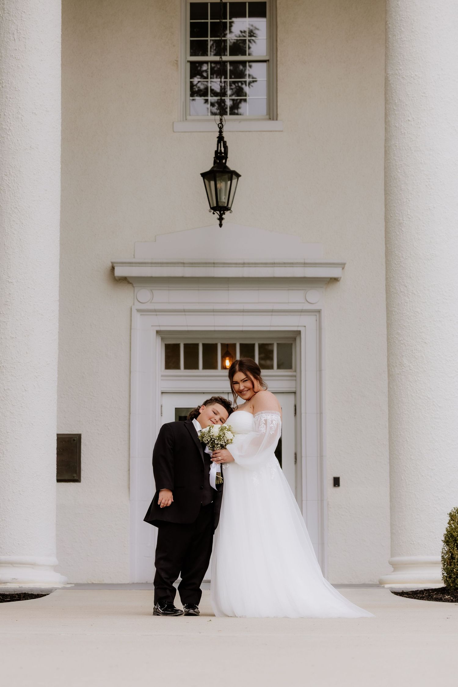 Bride in white gown and groom in black tuxedo pose together holding bouquet outside elegant white building.