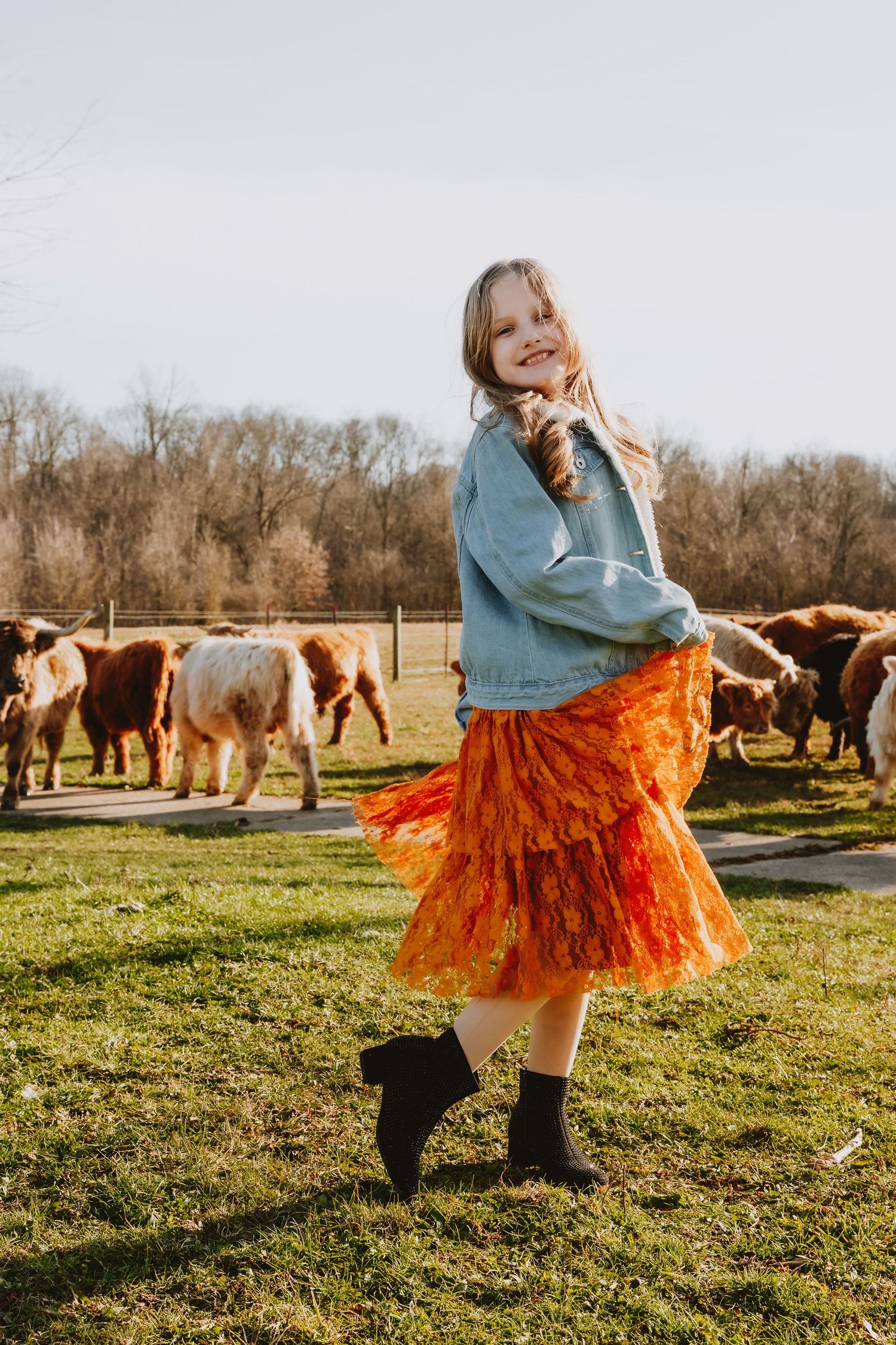 Young girl in orange ruffle skirt and denim jacket twirling in a field with Highland cattle grazing behind her.