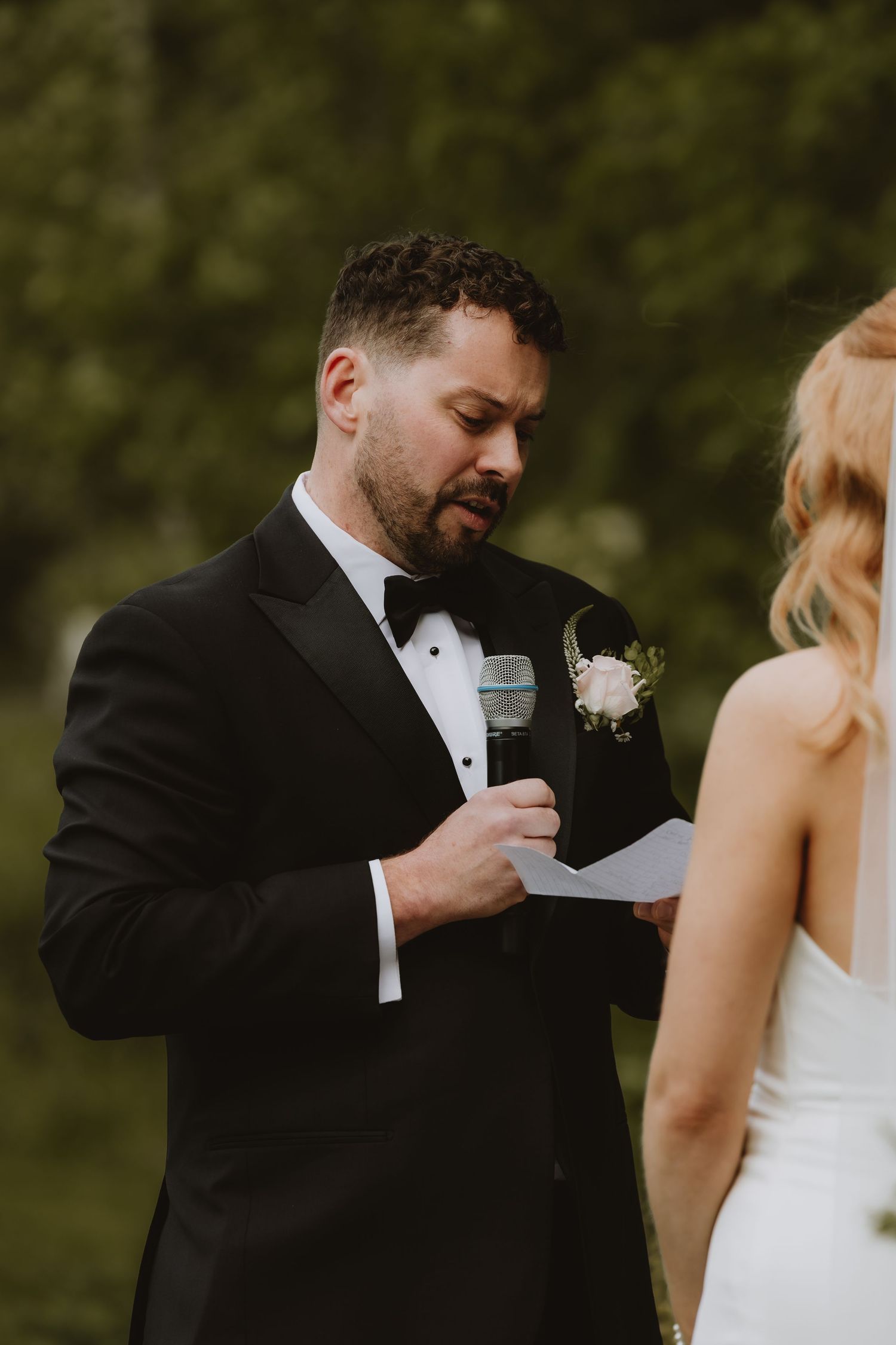 Groom in black tuxedo reads wedding vows from paper during outdoor ceremony, with bride in white dress facing him.