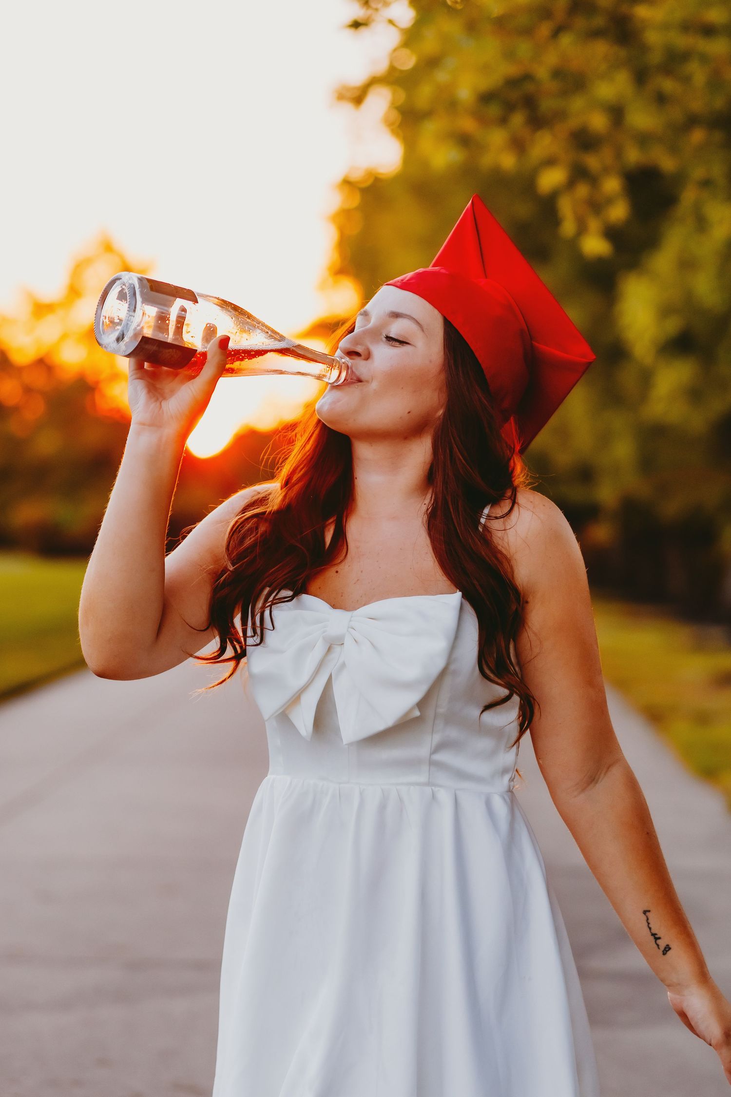 Person in white dress drinking from a bottle at sunset in a park setting.