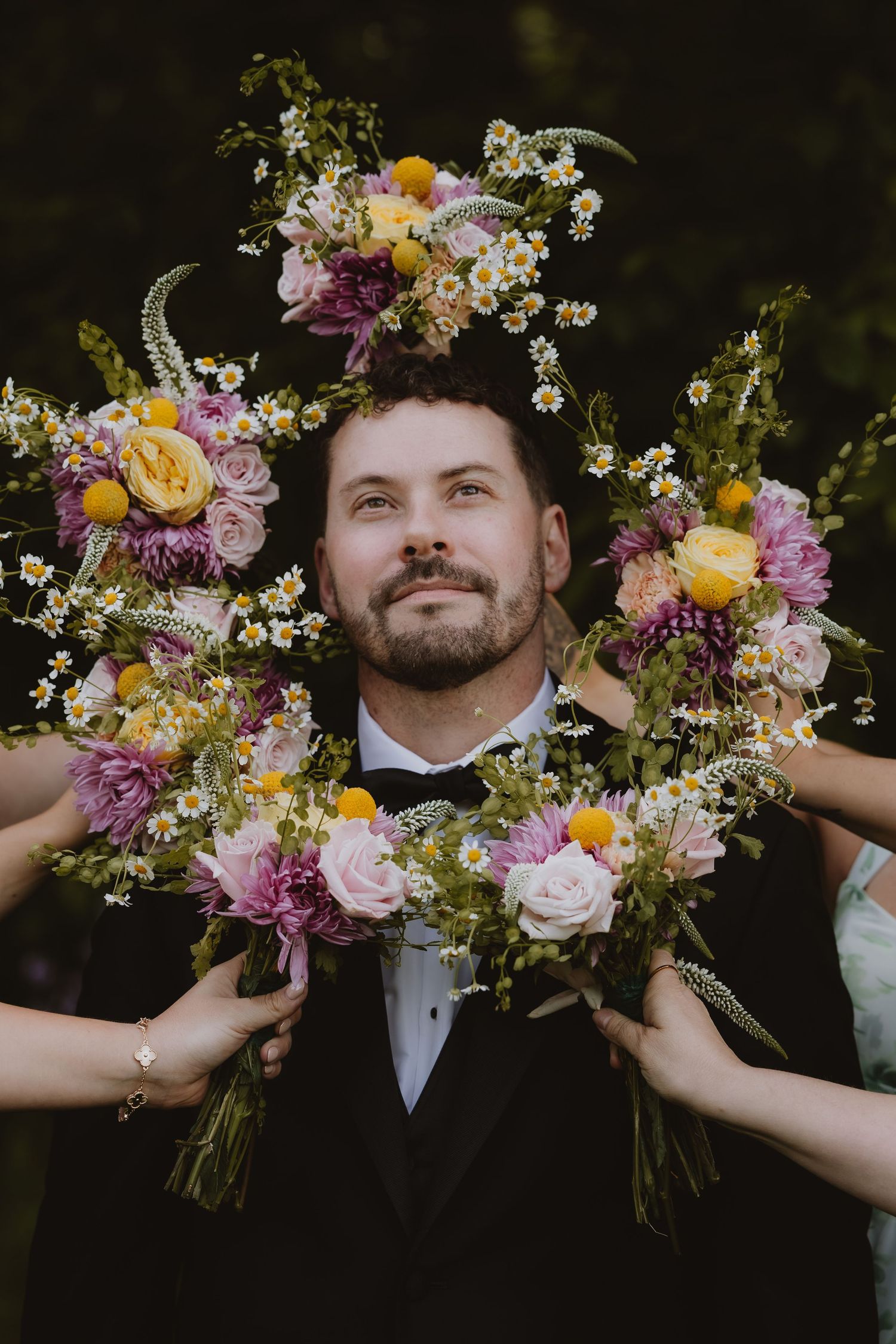 Groom in black tuxedo surrounded by colorful wildflower bouquets with pink roses, yellow blooms, and daisies at wedding.