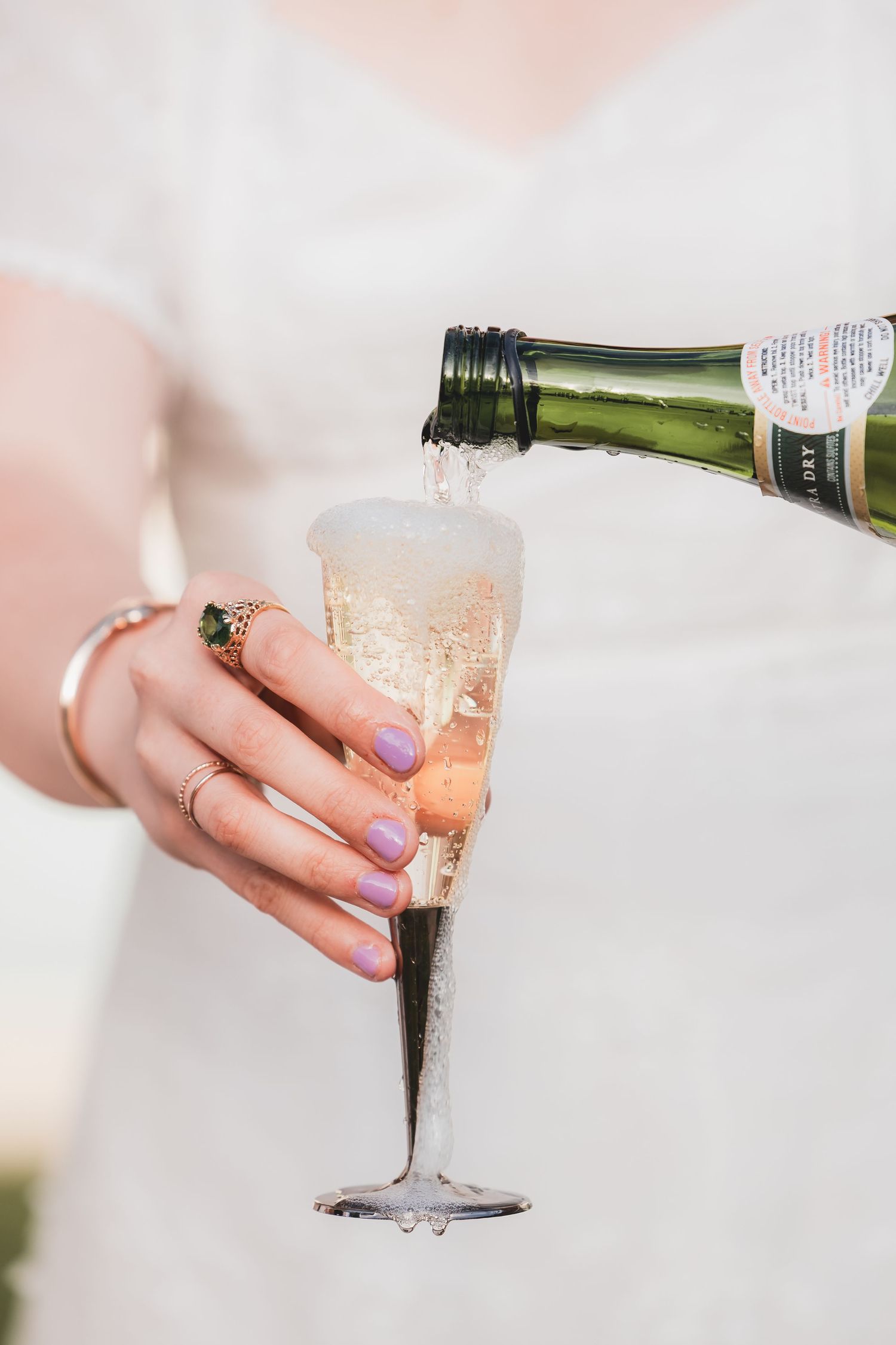 Bride in white dress pouring champagne into a flute glass, wearing purple nail polish and a gold ring.