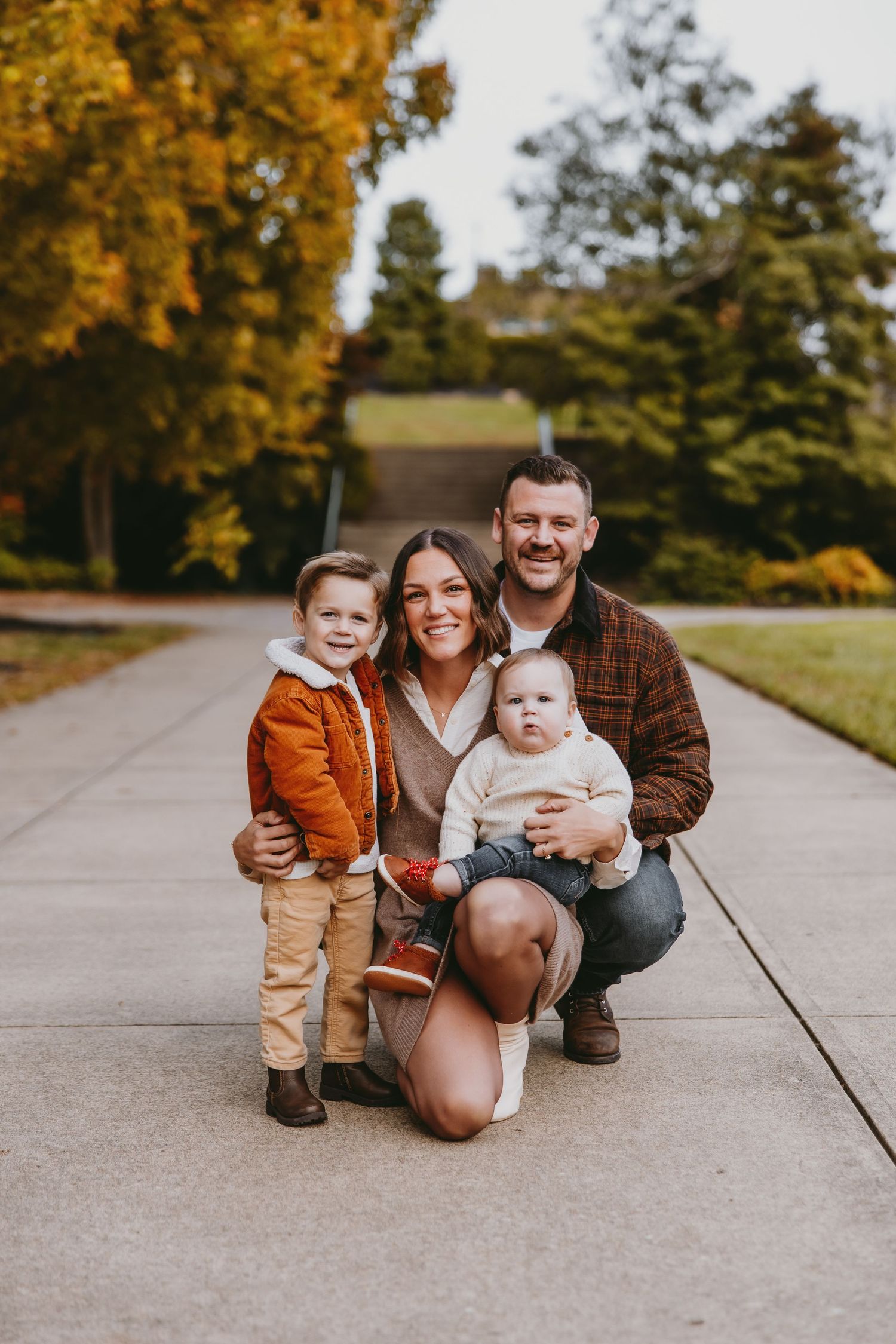 Family of four posing outdoors in fall, parents with toddler and baby on a pathway surrounded by autumn trees.