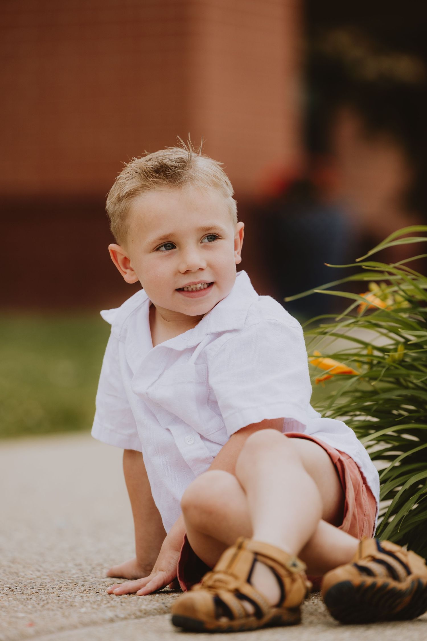 A young blonde boy in a white shirt and red shorts sits smiling outdoors near green plants in sandals.