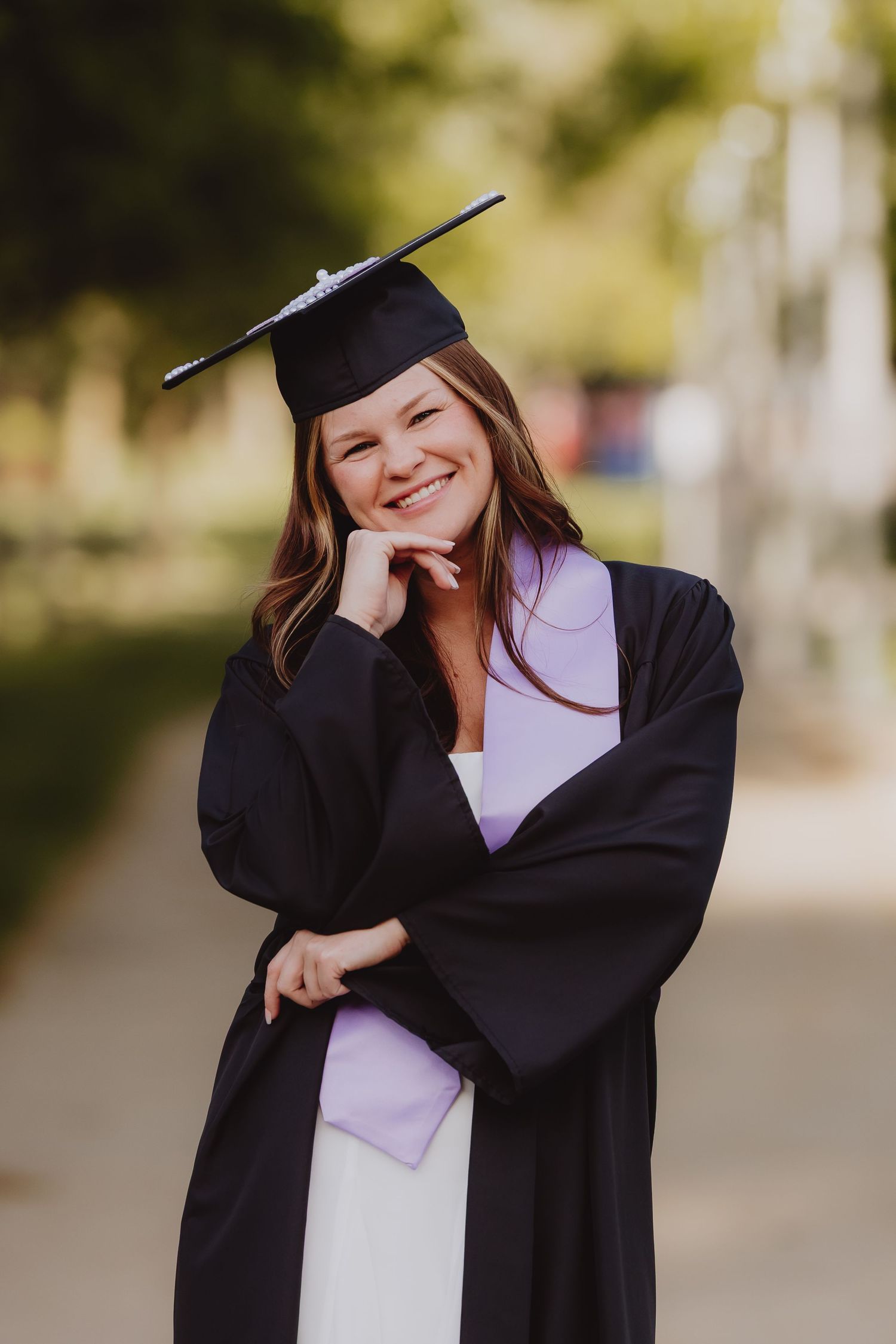 Smiling young woman in black graduation cap and gown posing outdoors with hand on chin on a blurred green background.