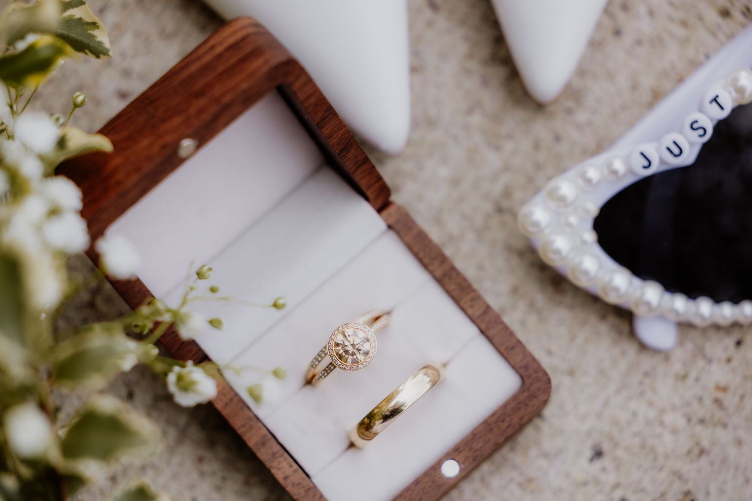 Gold engagement ring and wedding band displayed in a wooden ring box, with white flowers and bridal shoes nearby.