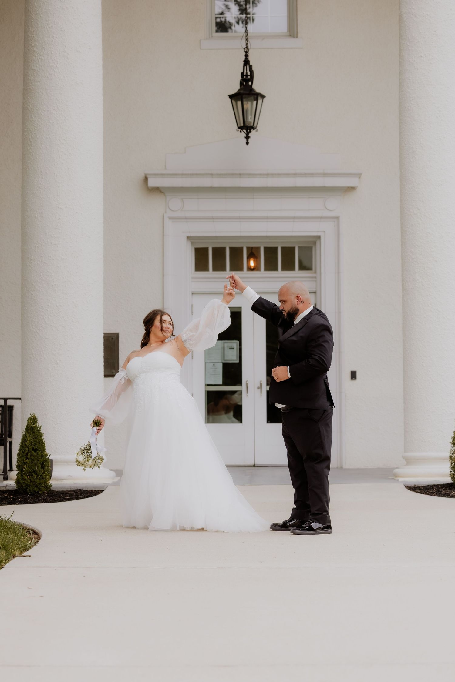 Bride in white tulle gown and groom in black suit dancing outside a white columned wedding venue entrance.