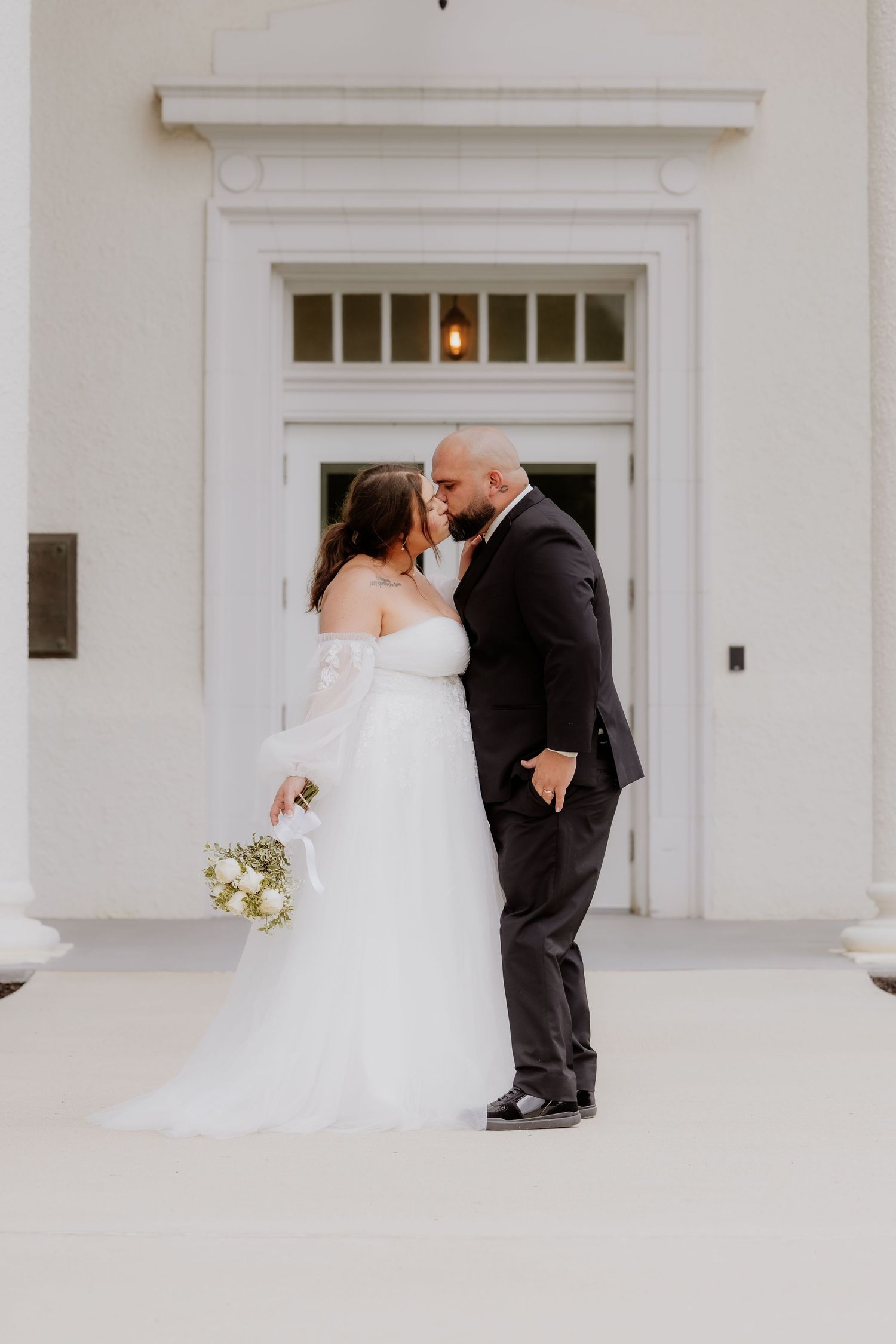 Bride and groom share a kiss outside a white building, bride holding a bouquet in an off-shoulder gown.