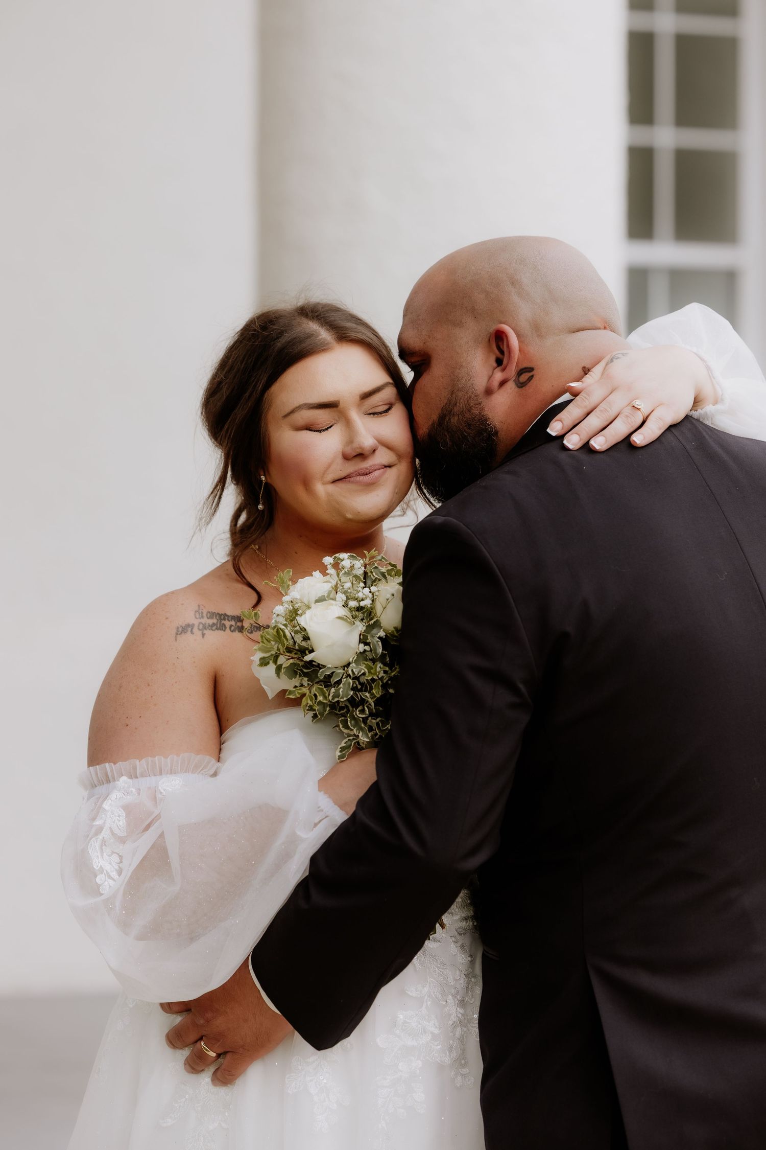 Bride in white off-shoulder gown holds bouquet while groom in black suit embraces her tenderly outdoors.