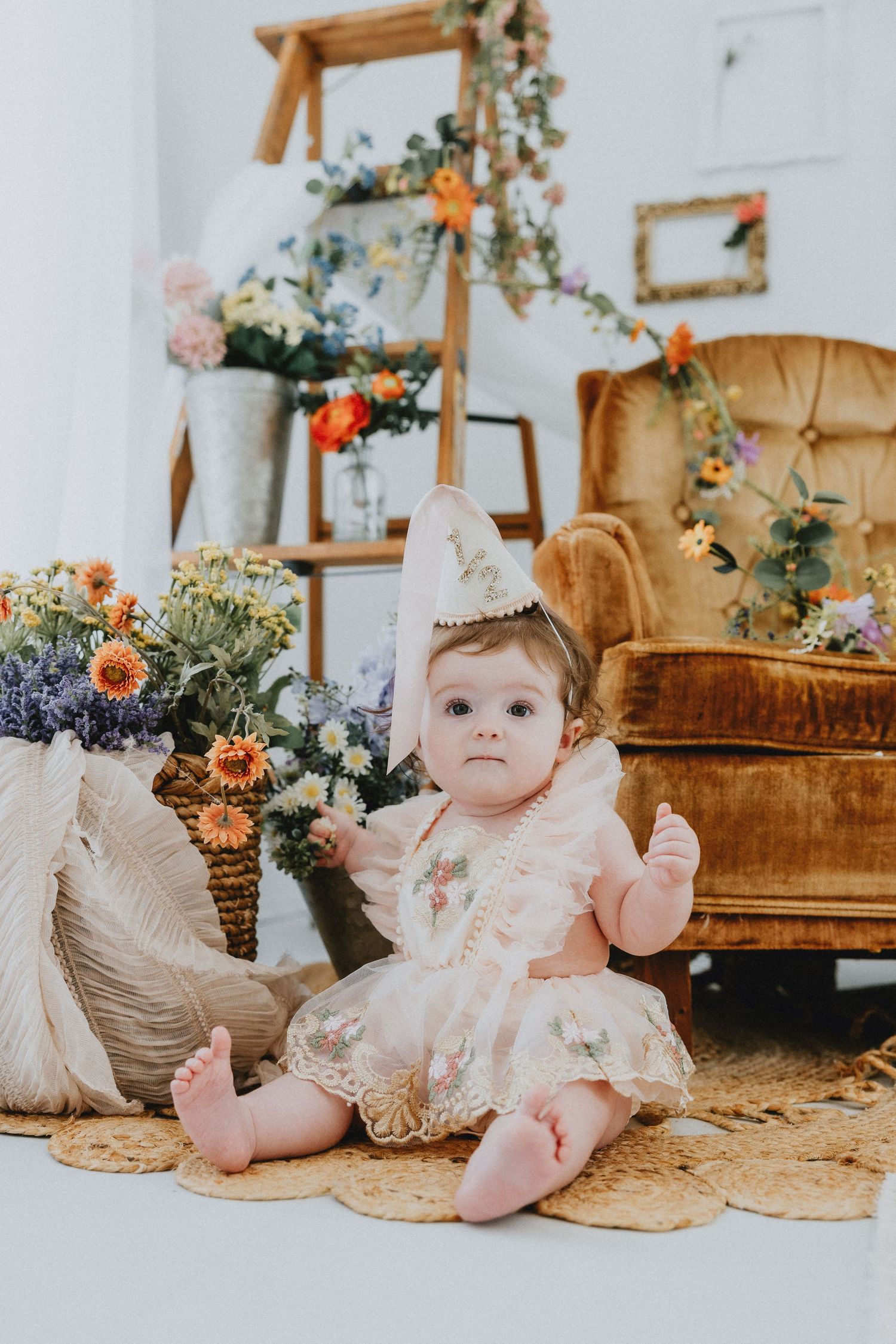 A baby girl in a floral dress and party hat sits on a rug surrounded by colorful flowers and a velvet chair.