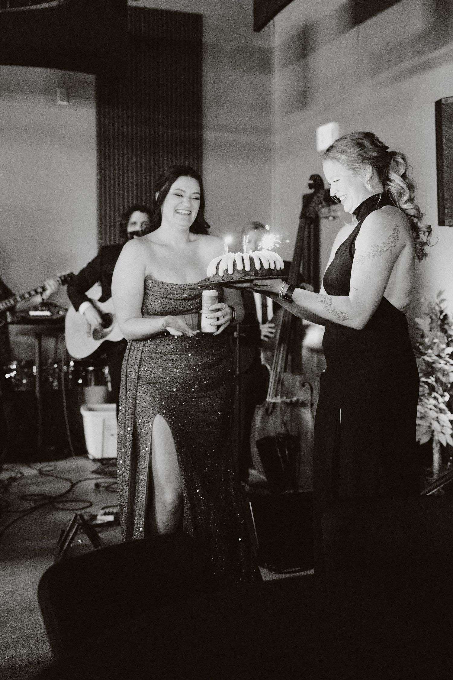 Black and white photo of two women sharing a cake at a formal event with live band performing in background.